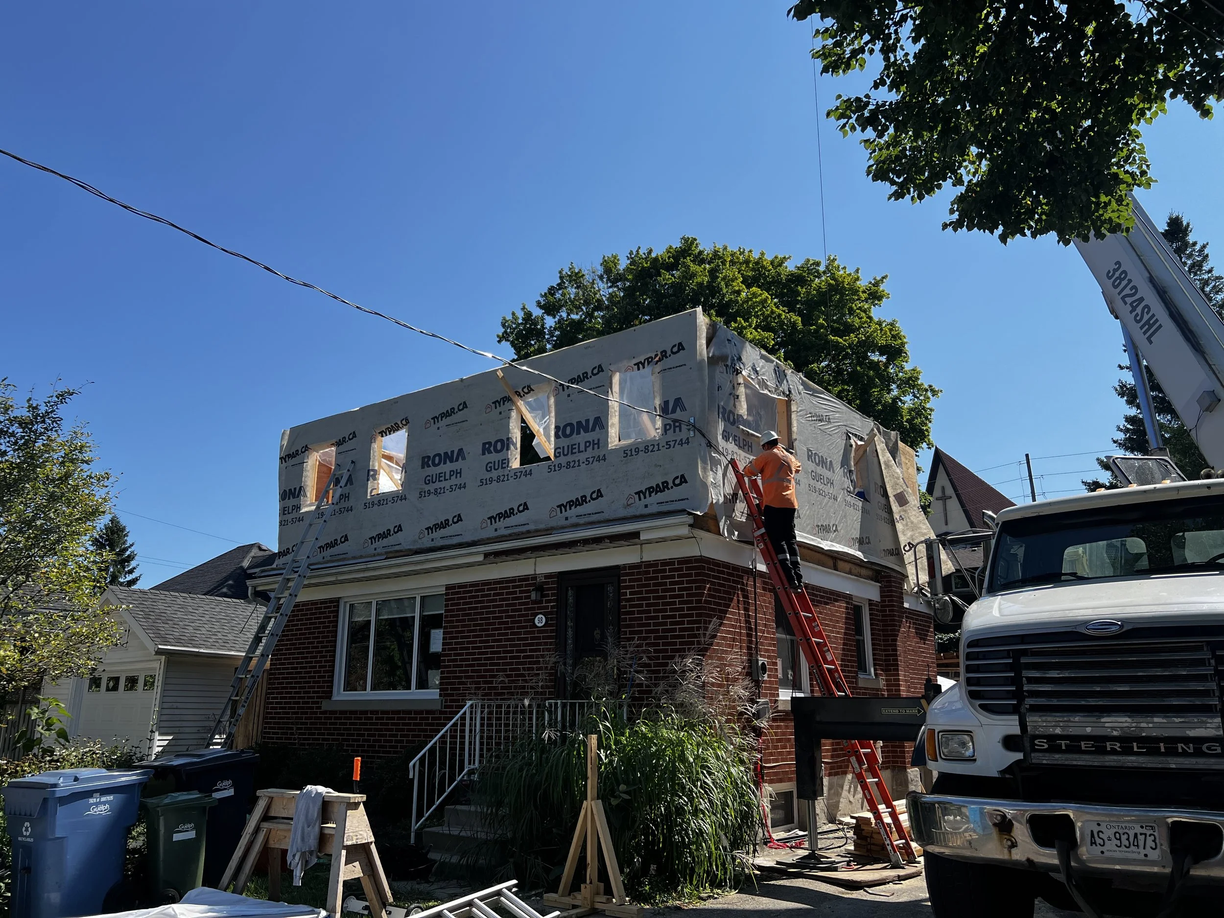 Construction workers installing new roofing on a brick house with a crane, ladders, and construction materials outside on a sunny day.