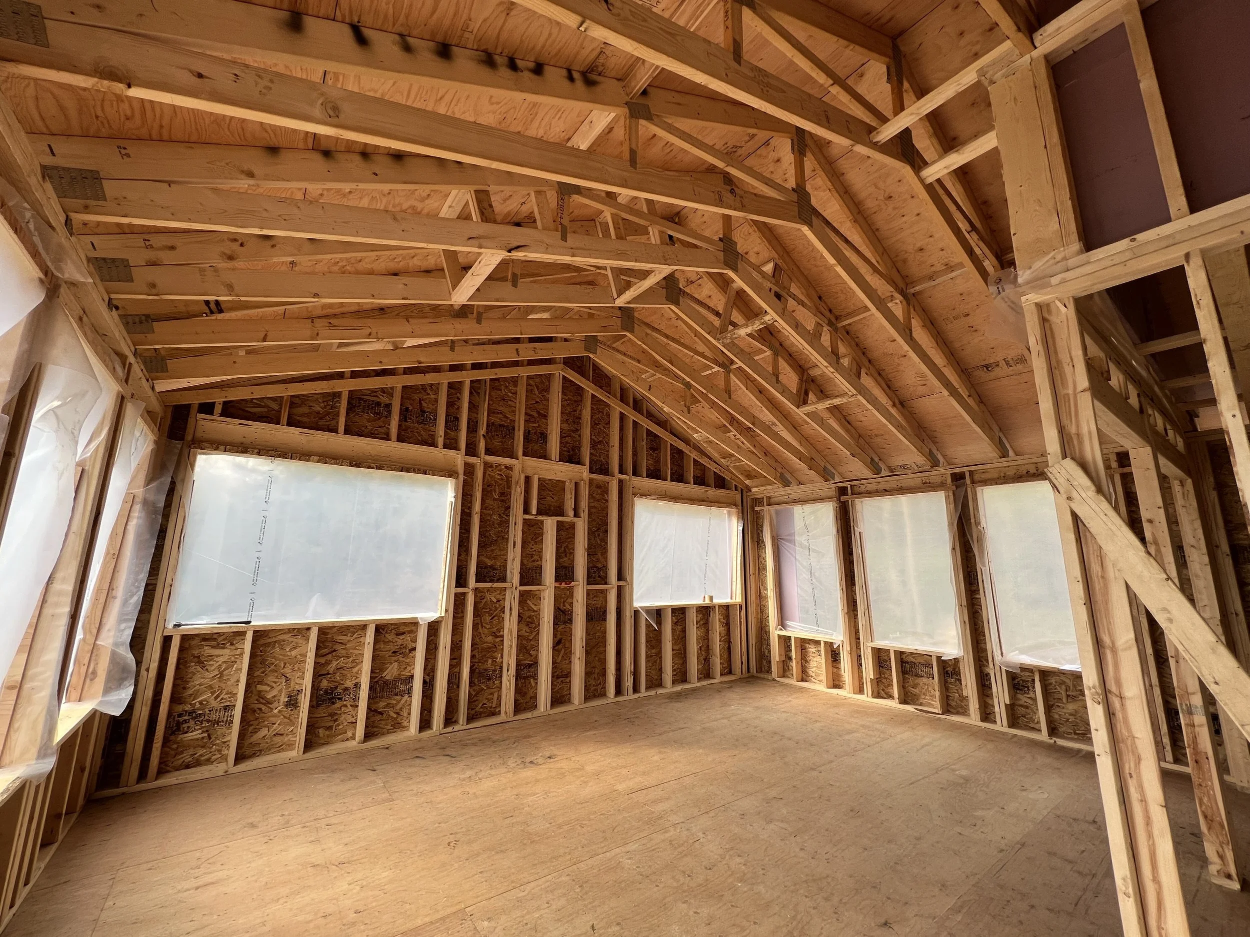 Interior view of a house under construction, showing wooden framing and exposed plywood walls and ceiling, with plastic-covered window openings.