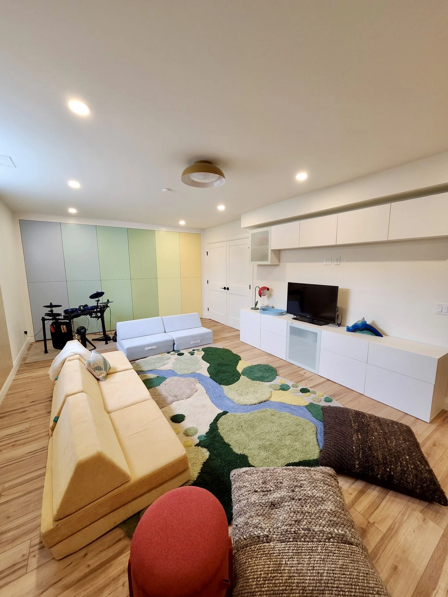 Modern living room with yellow sofa, patterned carpet, white cabinets, TV, colorful wall panels, upholstered armchair, throw pillows, exercise bike, and wooden flooring.