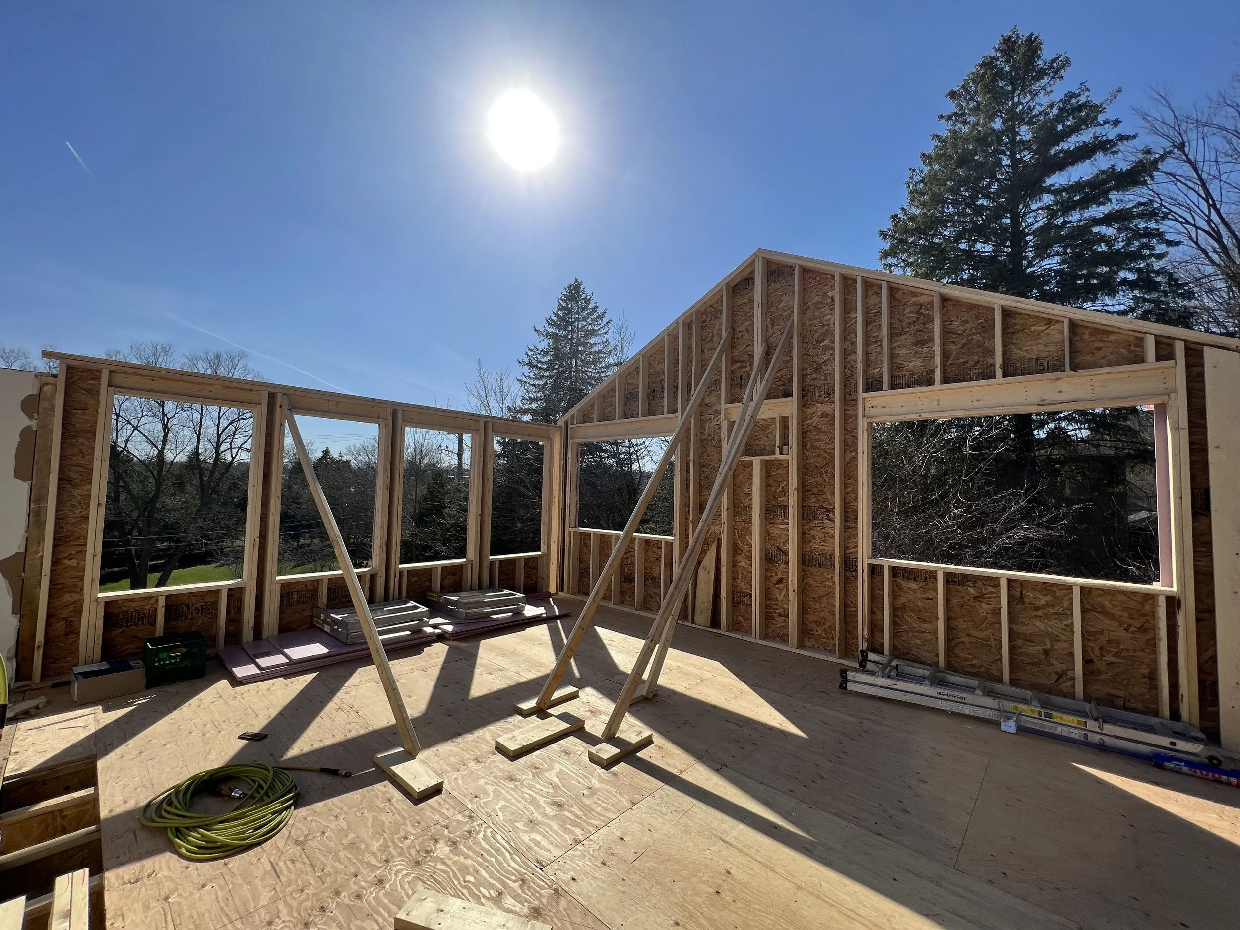 Wooden house frame under construction with open walls, exposed beams, and a clear blue sky in the background. Sun is shining brightly, creating distinct shadows.