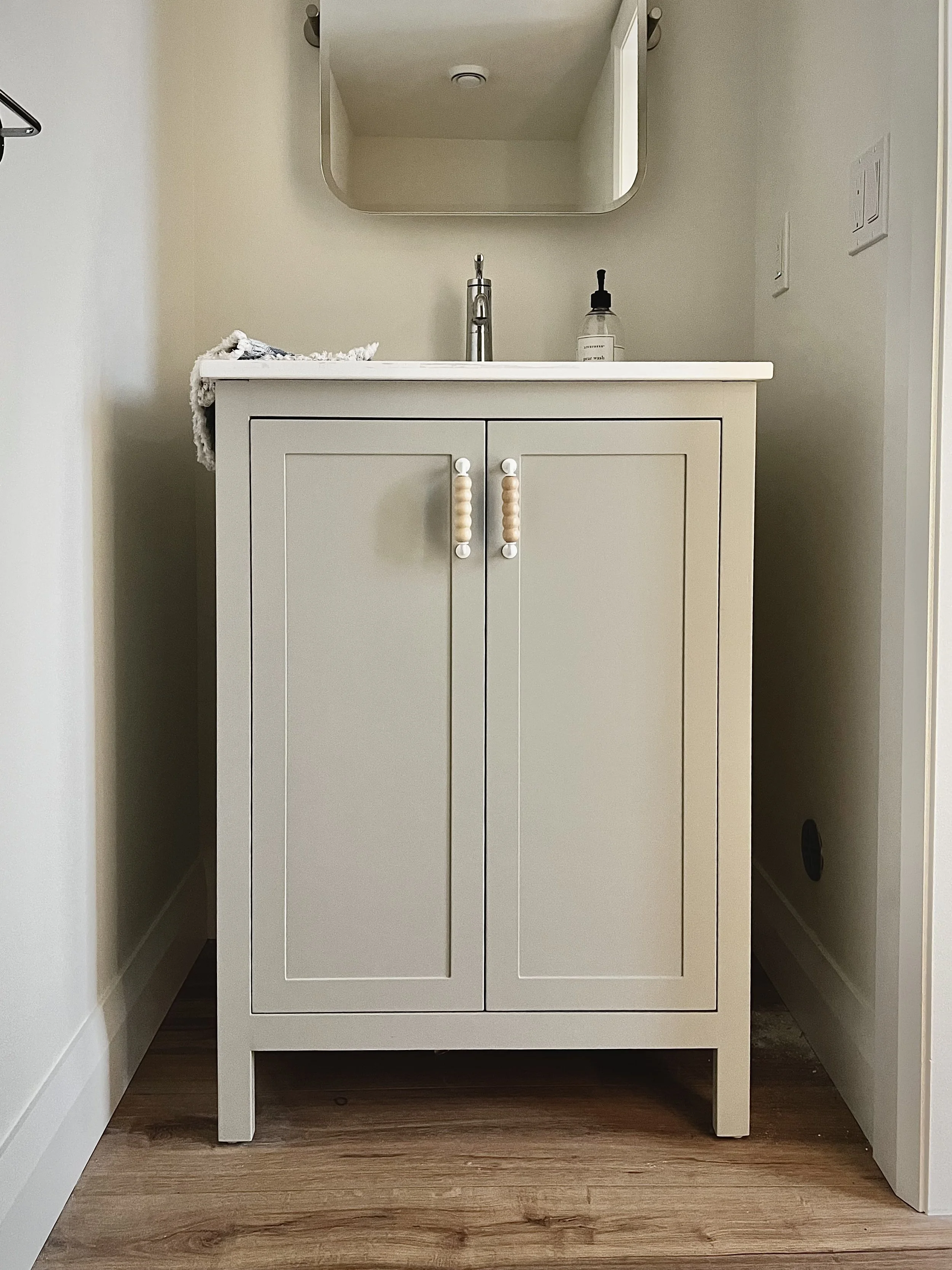 White bathroom vanity with two cabinet doors and round bead handles, a silver faucet, a glass soap dispenser, and a mirror above it, set against beige walls and wooden flooring.