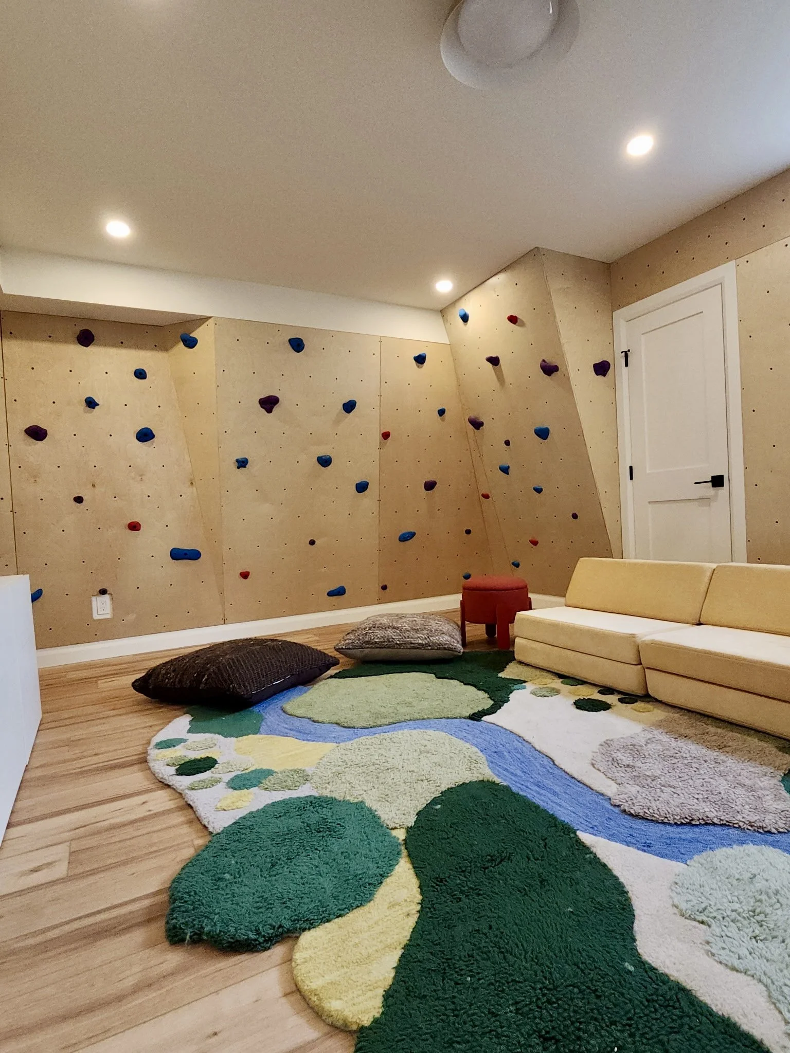 Indoor room with a textured climbing wall featuring colorful holds, a beige sofa, a red stool, several pillows on a decorative rug with a geometric pattern, and a white door.
