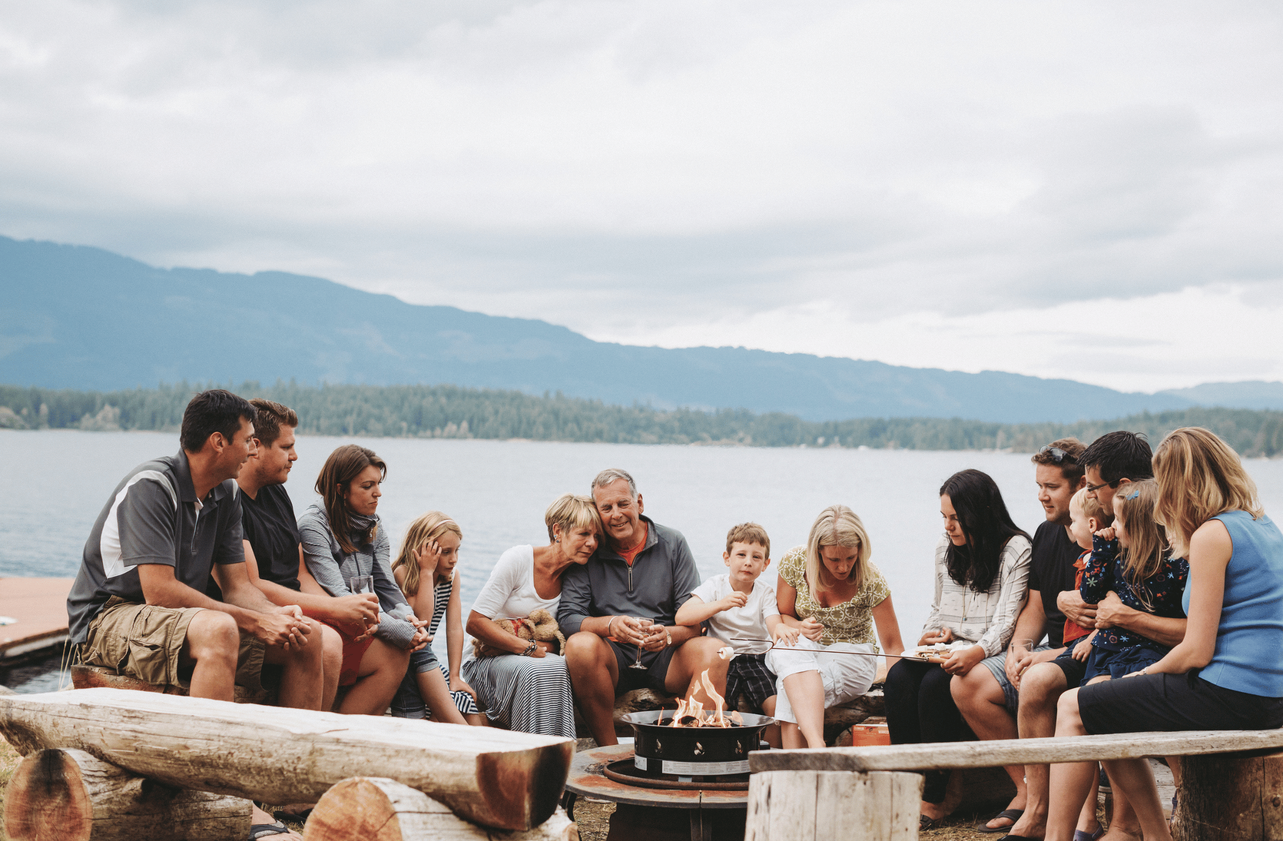 Family and friends gathered around a fire pit on a lakeside outdoor patio, with mountains in the background.