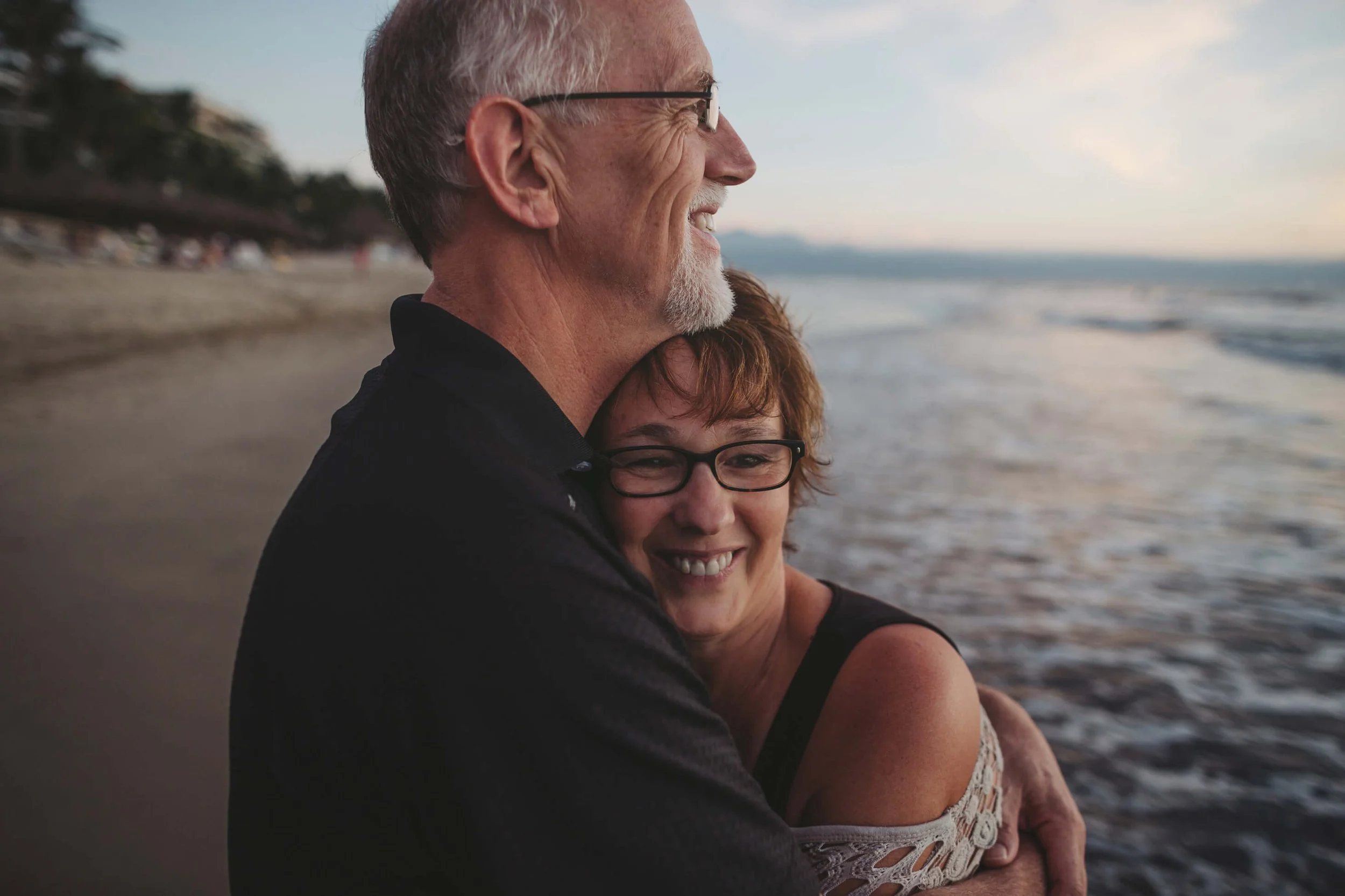 An elderly man and woman hugging on a beach at sunset, both smiling and wearing glasses.