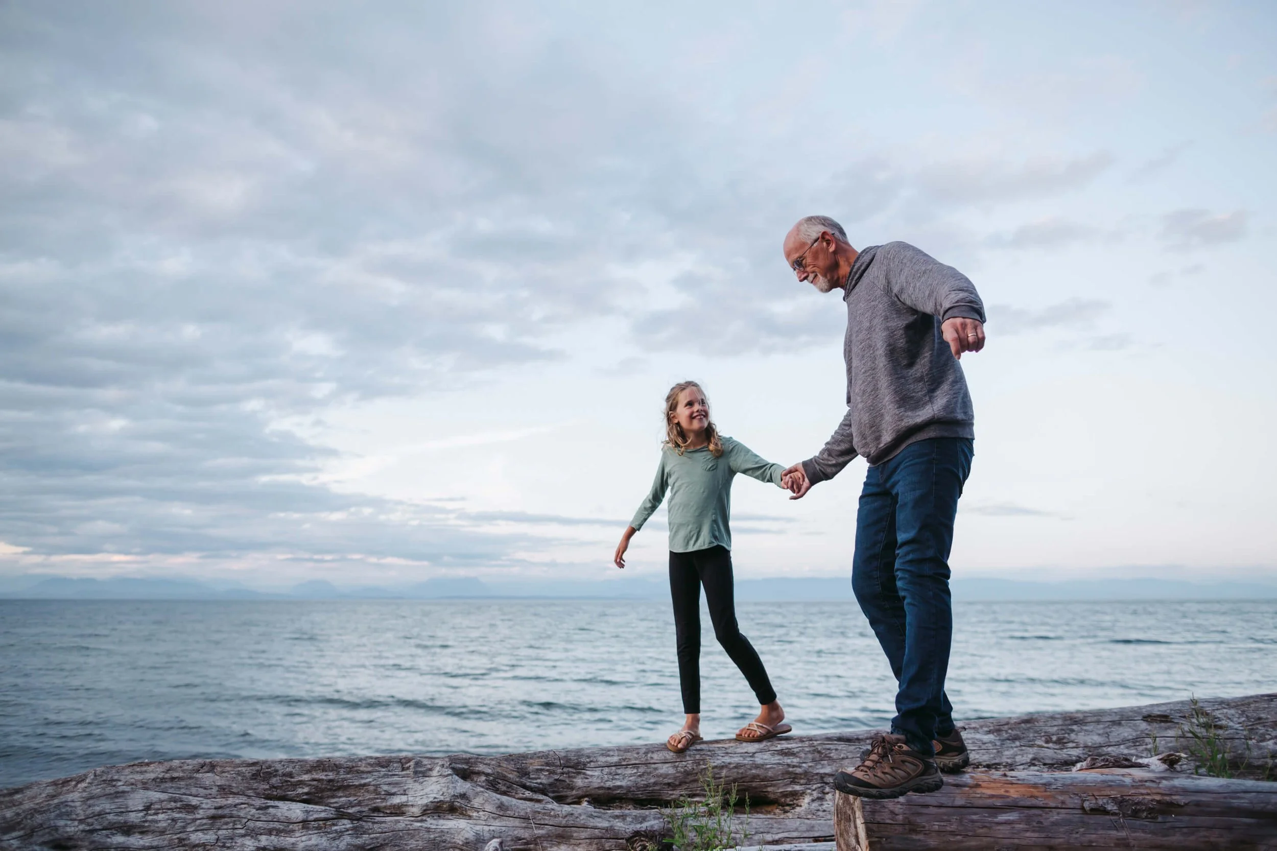 A girl and an elderly man playing together on a log by the water on a cloudy day, holding hands and smiling.