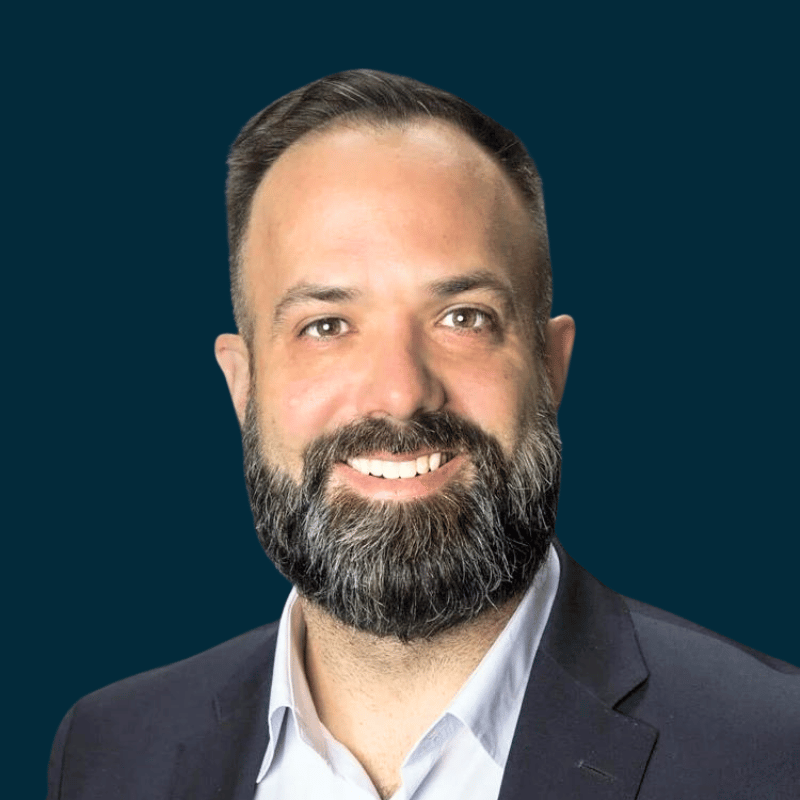 Portrait of a man with a beard and short hair, wearing a suit and white shirt, smiling against a dark background.