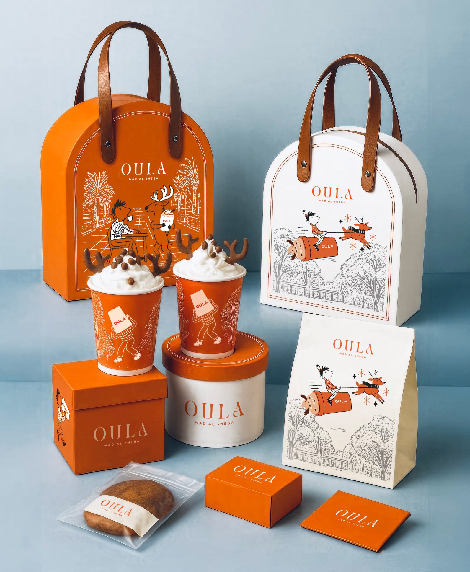A collection of bakery and packaging items, including orange and white boxes and bags with the logo 'OULA,' two cups of whipped cream topped with mini chocolate chips and reindeer antlers, a cake or bread wrapped in clear plastic, and decorative illustrations of people and reindeers on the packaging, set against a light blue background.