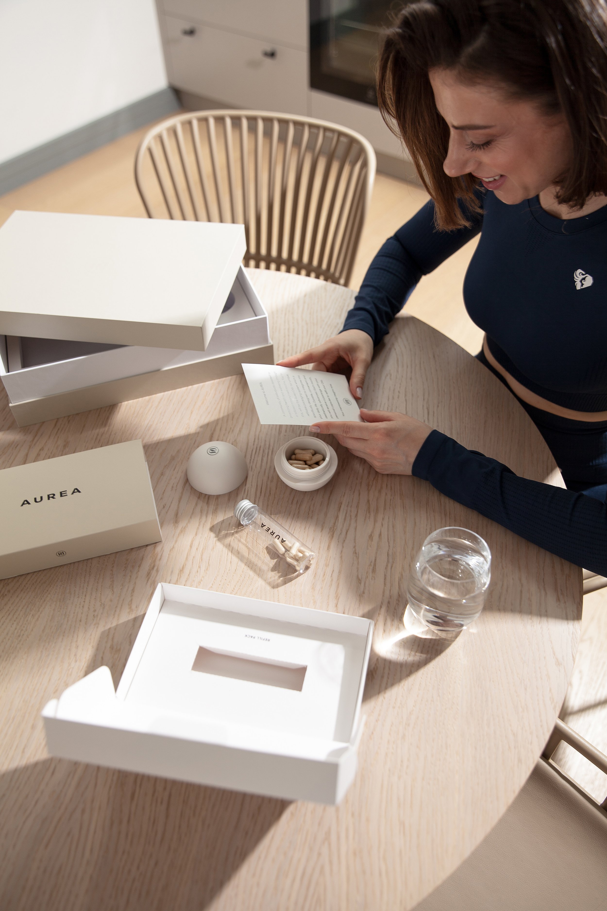A woman with short brown hair wearing a navy blue long sleeve shirt is looking at a small card on a light wood table. There are several empty white boxes and containers, a glass of water, and a clear glass ampoule on the table, in a room with a papered wall and a kitchen cabinet in the background.