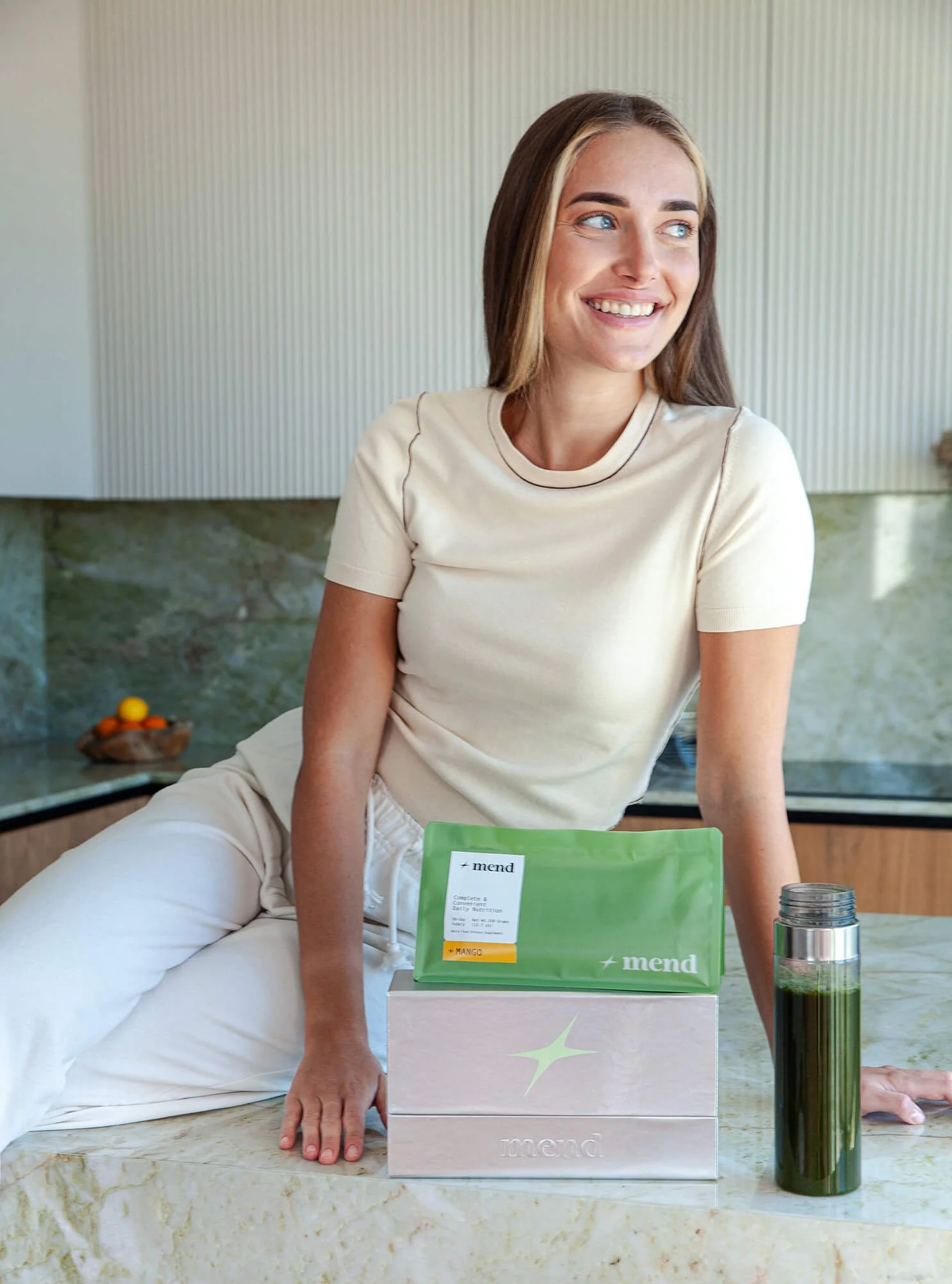 A woman sits at a marble countertop with a green and white package labeled 'mend' in front of her. She is smiling and looking to the side, wearing a beige t-shirt and white pants, with a silver thermos and a glass bottle beside her.