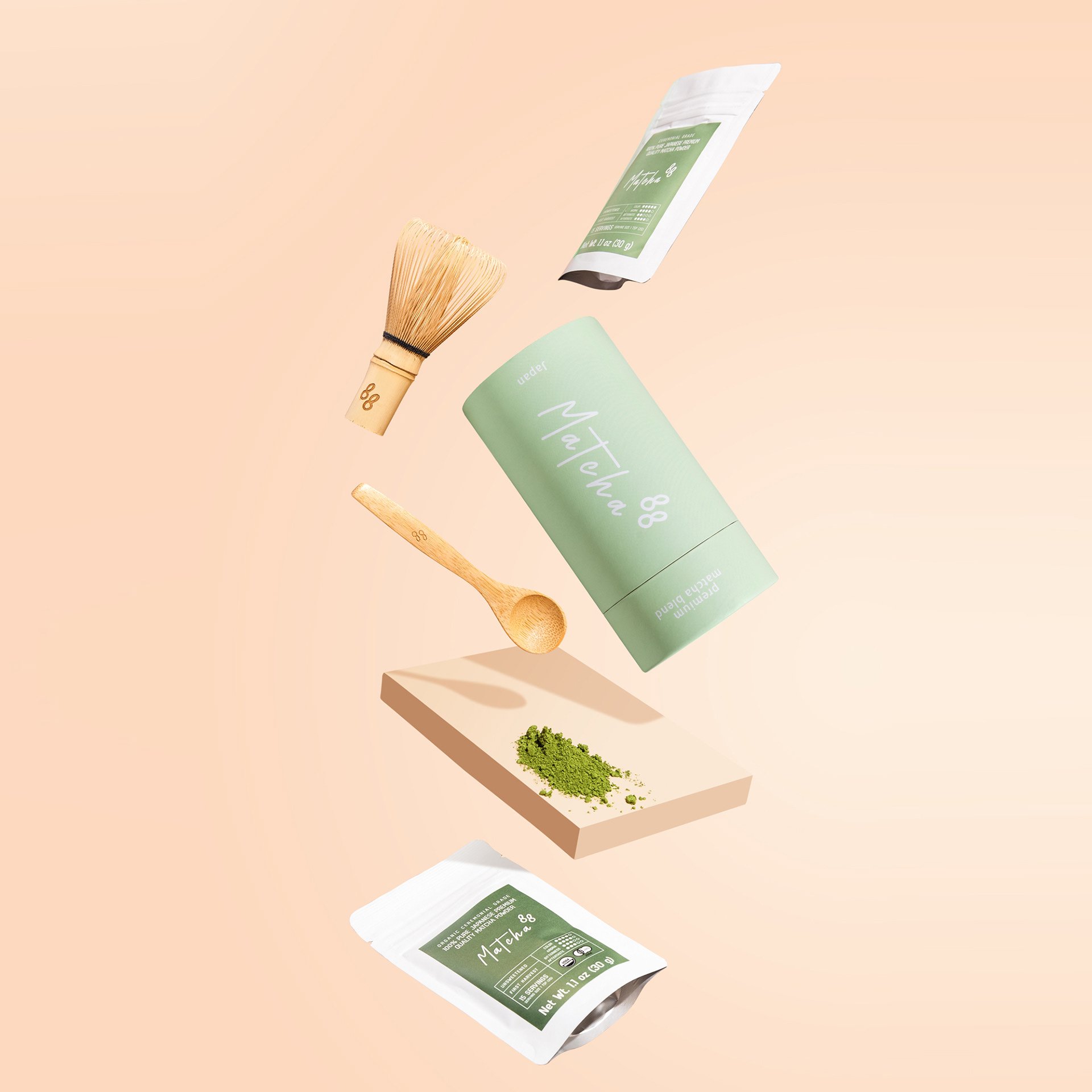 A green package labeled "Matcha" with a bamboo whisk and a wooden spoon with the number 88, and a small pile of green matcha powder on a beige platform against a peach background.