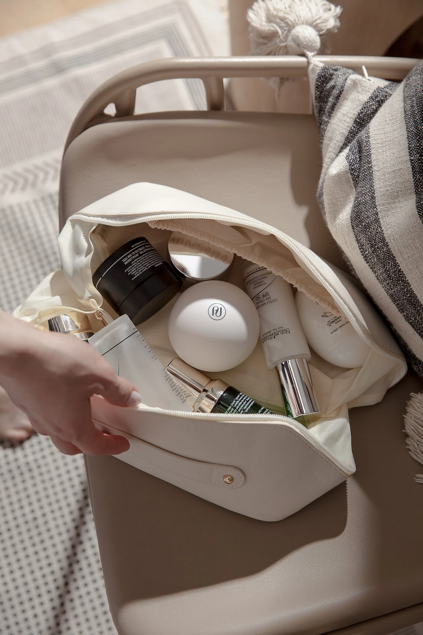 A white handbag with a zipper open, containing assorted skincare and beauty products, placed on a beige surface. A hand is seen holding the bag open.