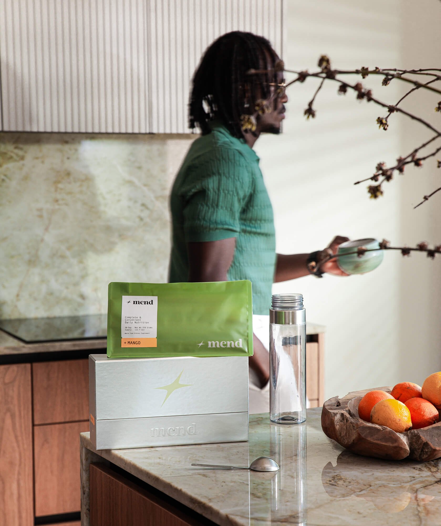 Man with dreadlocks wearing a green shirt standing behind a countertop, holding a mug, with a piece of paper and fruit on the counter, in a home kitchen setting.