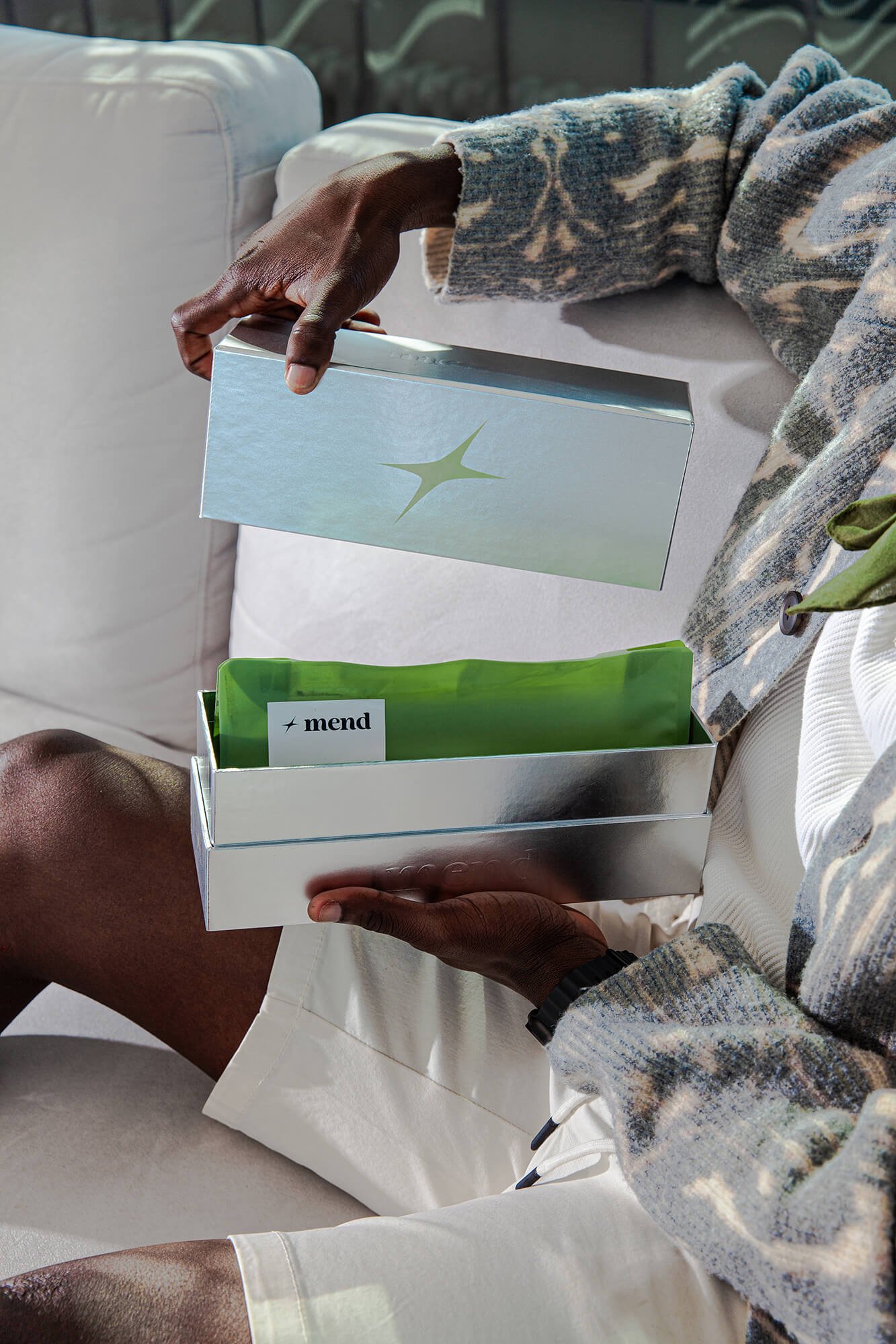 Person with dark skin holding a white box with a green star logo, sitting on a white couch with a patterned sweater and white pants, with another person with dark skin sitting close by.