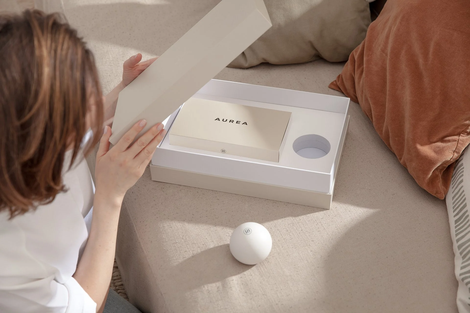A woman with reddish hair examining a box labeled 'AUREA' inside a white box with a circular slot, on a beige couch with pillows.