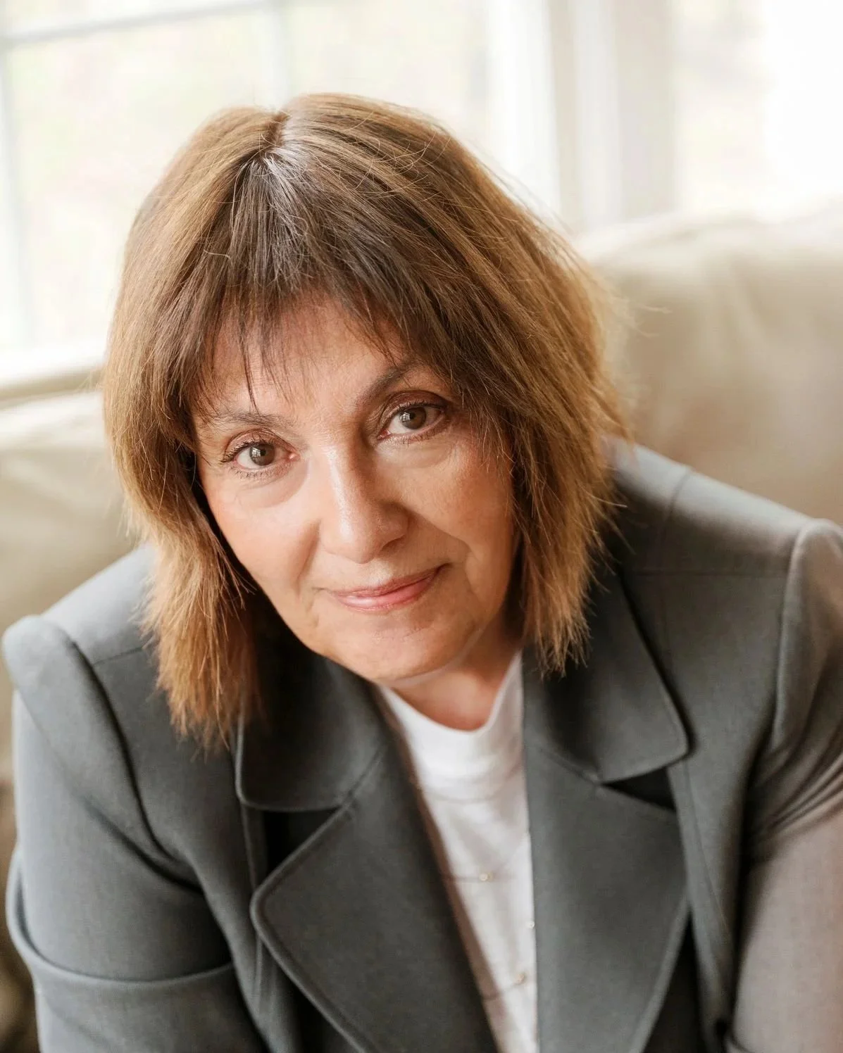 A woman with shoulder-length brown hair, wearing a gray jacket and white shirt, sitting on a beige couch near a window, looking at the camera.