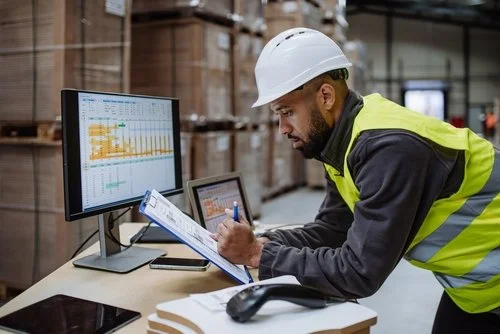 A man writing on a clipboard in front of a computer screen in a warehouse with stacked pallets in the background.