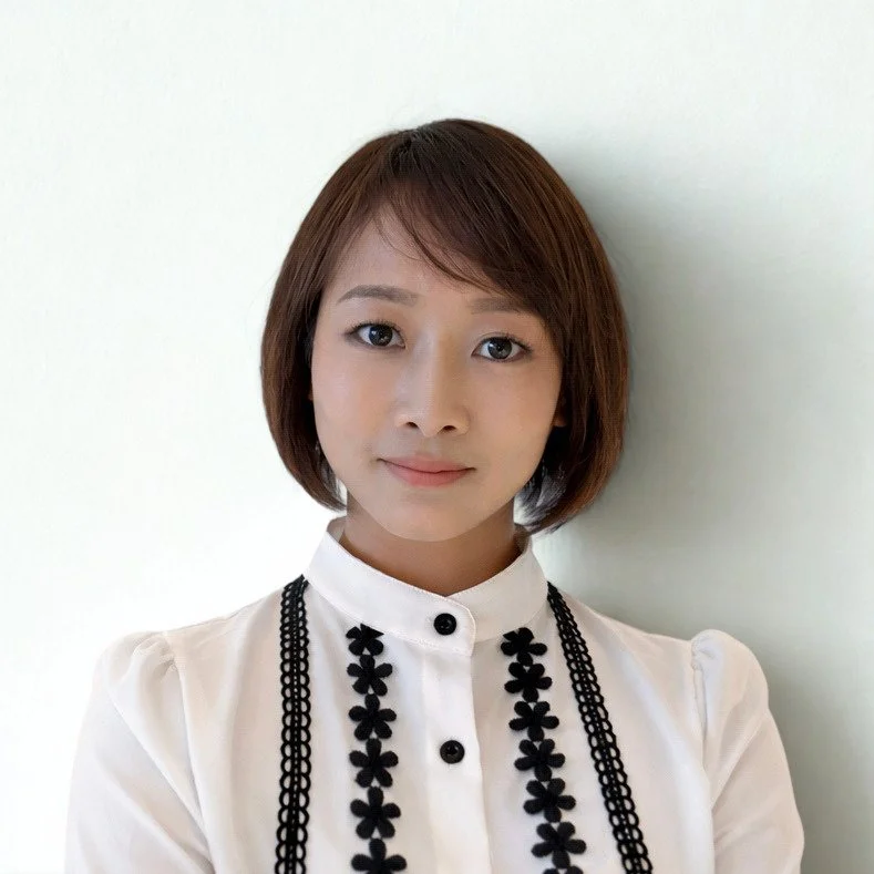 A woman with short brown hair wearing a white blouse with black embroidered floral designs, standing against a white wall.