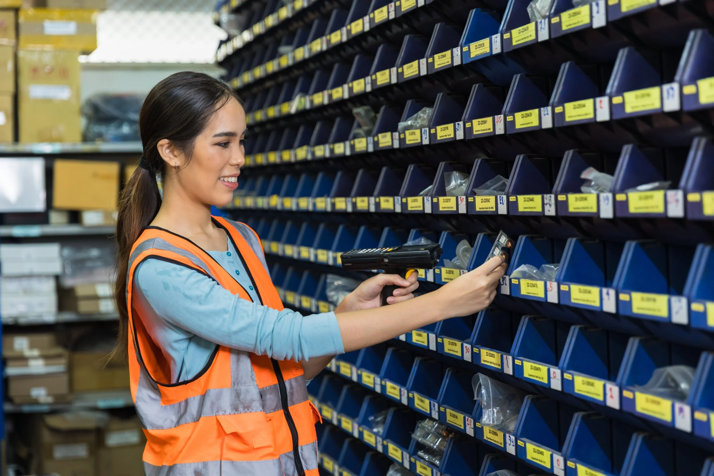 A female with brown hair and holding a scan gun, scanning small parts in blue storage bins.