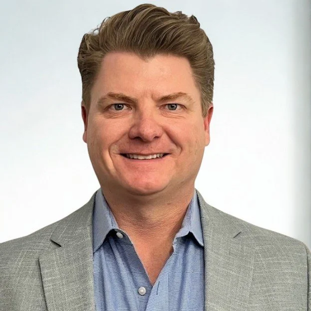 A man with light skin, brown hair, and blue eyes in a gray suit and blue shirt smiling at the camera.