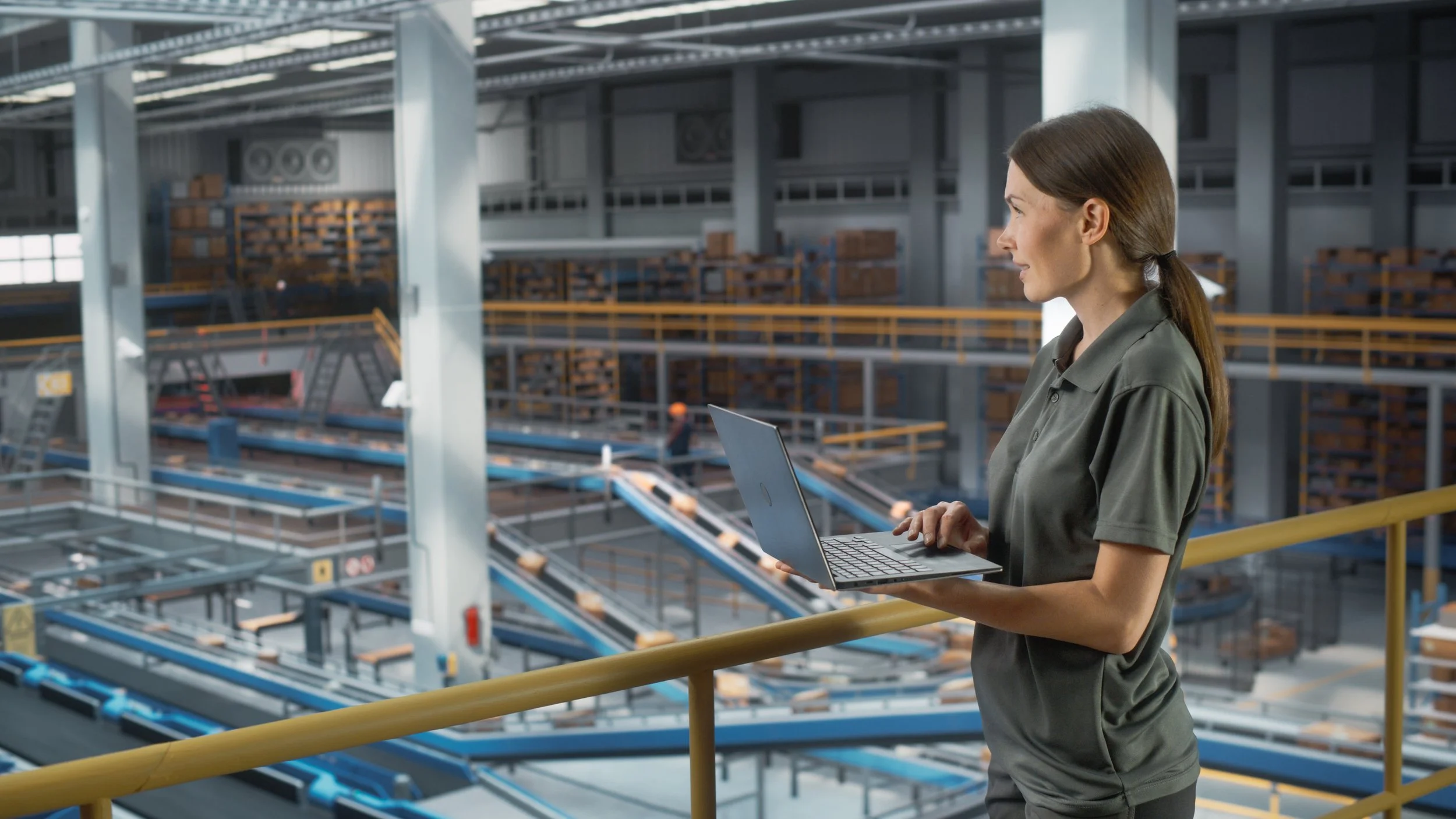 A female with brown hair overlooking a distribution facility on a balcony while holding a laptop.