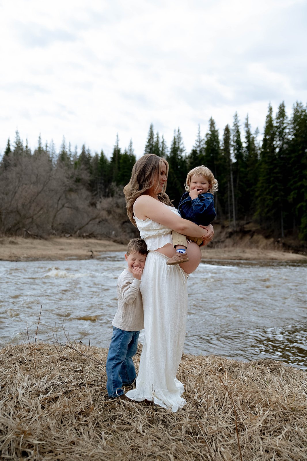 A woman holding a young boy while another boy stands beside her near a river, surrounded by trees with an overcast sky.