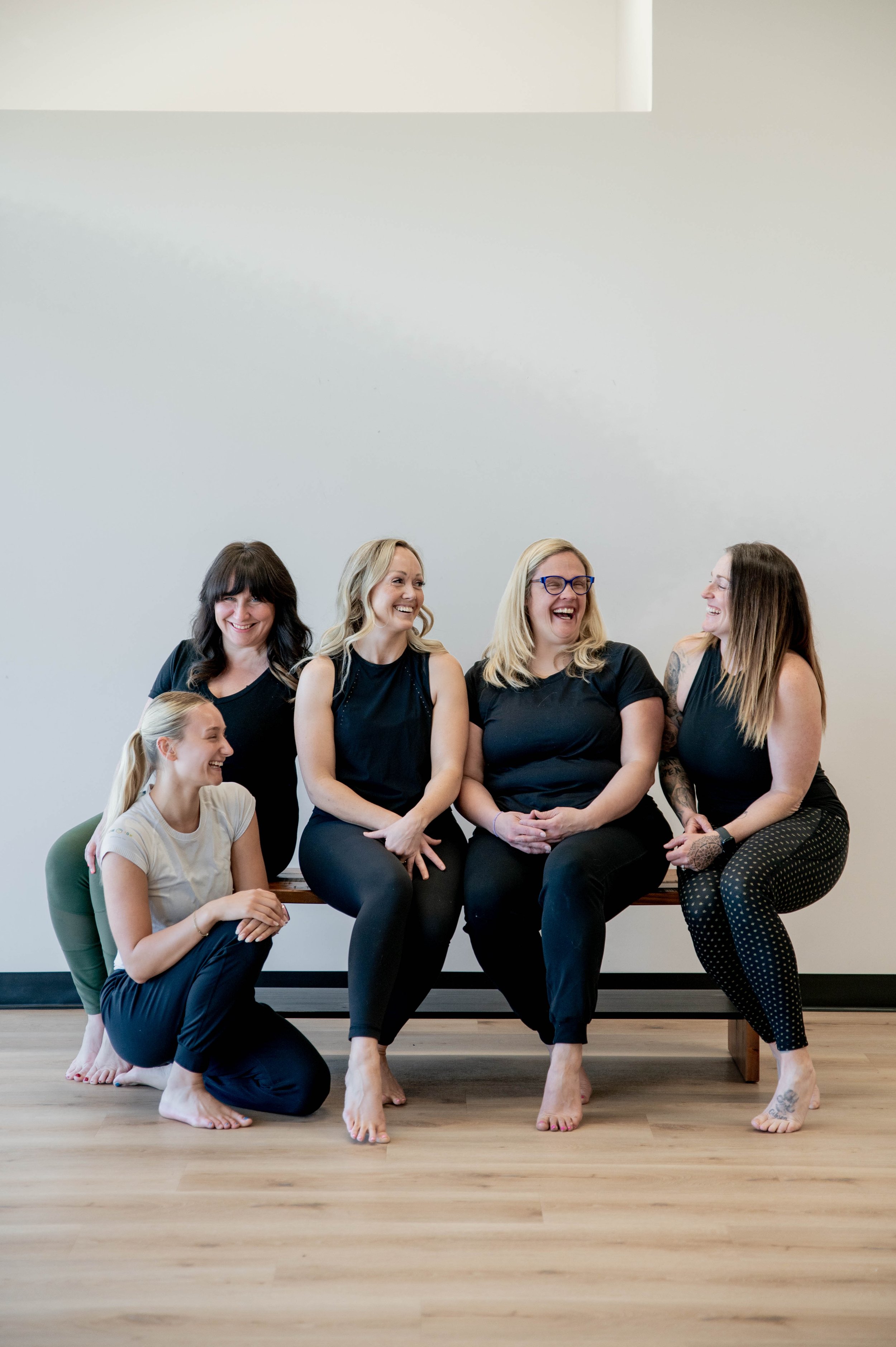 A group of six women laughing and smiling together, some sitting on a bench and others kneeling or standing, in a room with a wooden floor and a plain, light-colored wall.