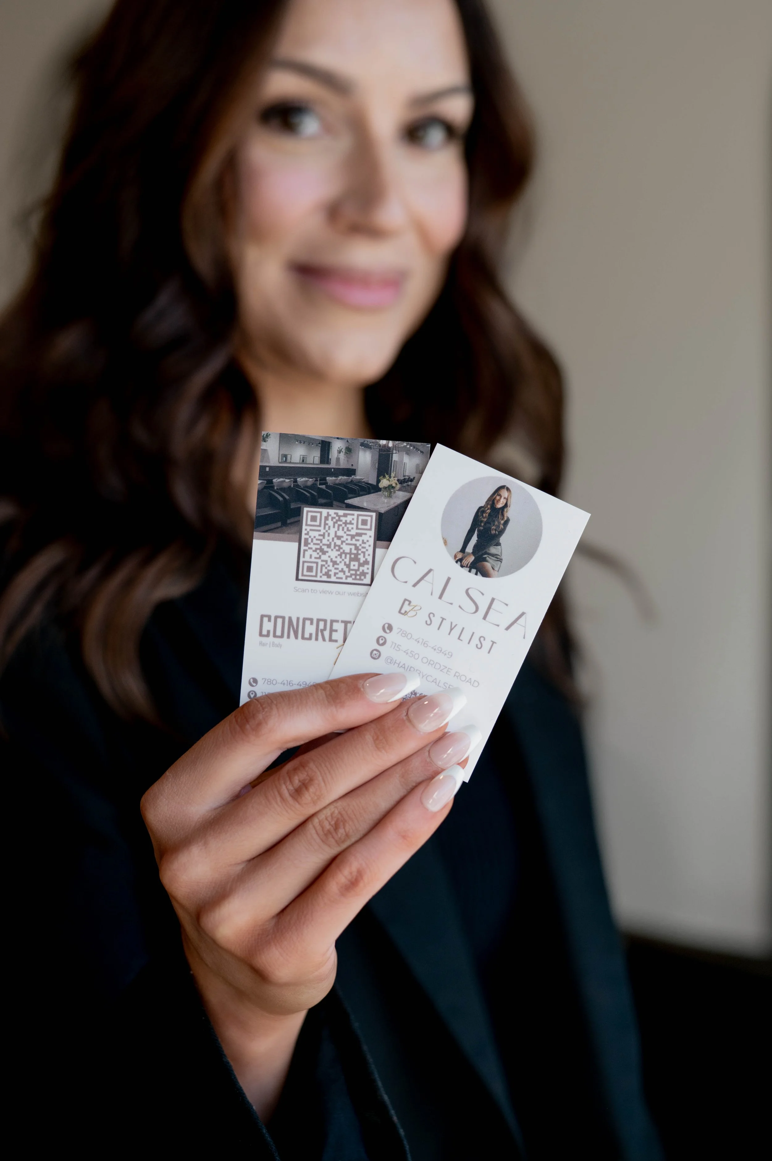 A woman with wavy brown hair holding business cards, one for a stylist named Calsea. She is smiling and wearing a dark blazer.