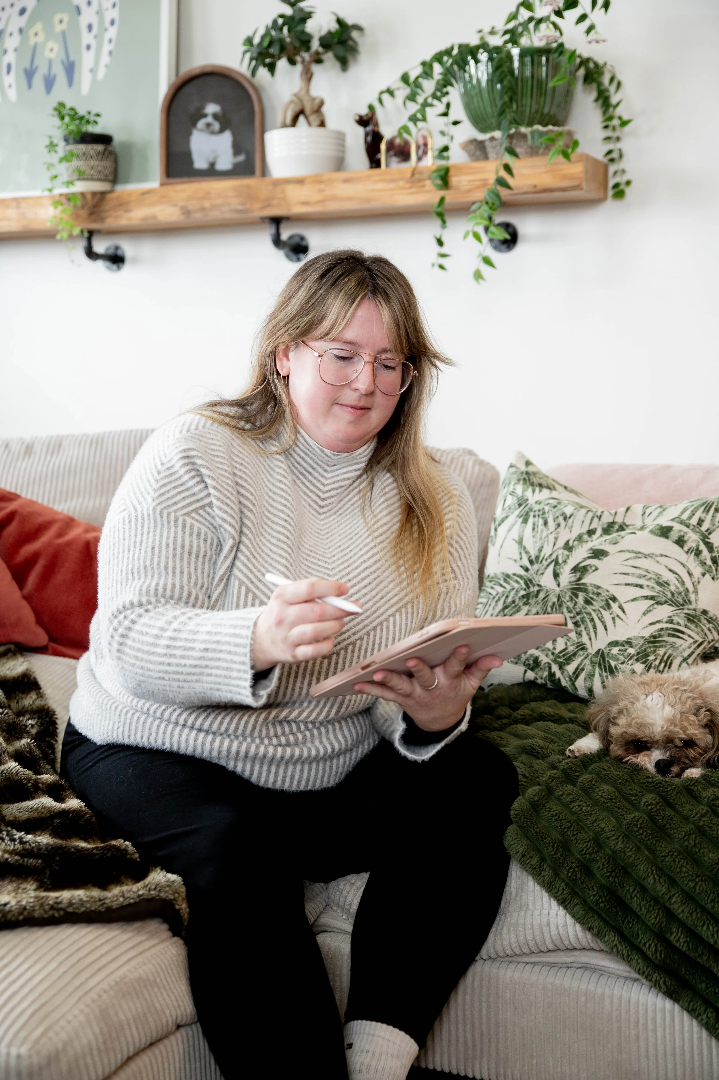 A woman with glasses and long hair sitting on a beige couch, holding a notepad and pen. She is wearing a striped sweater and black pants. A small dog is lying on a green blanket beside her. The background features a wooden shelf with plants and framed photos.
