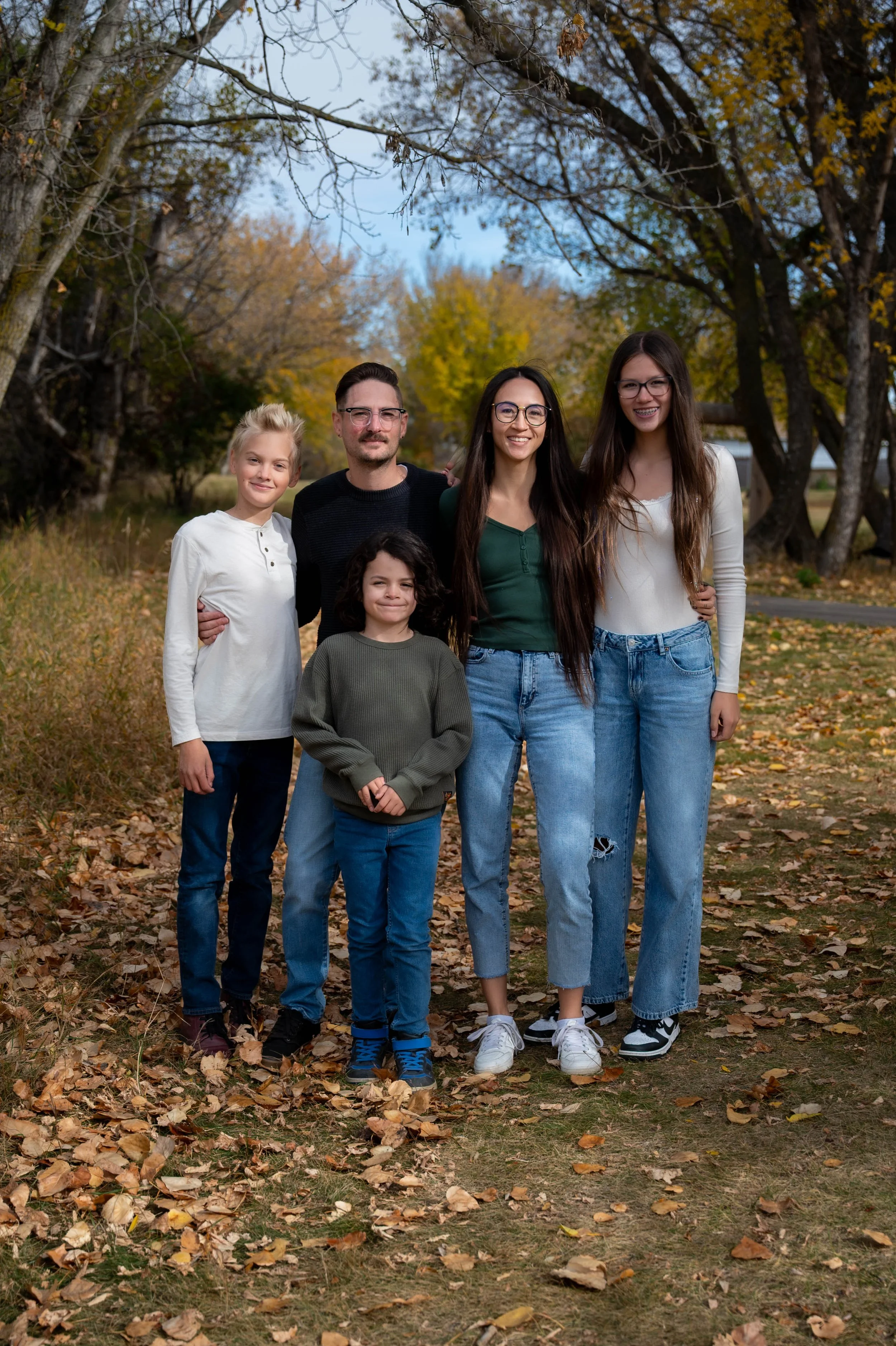 A family of five standing outdoors on a fall day among trees with colorful autumn leaves, smiling at the camera.