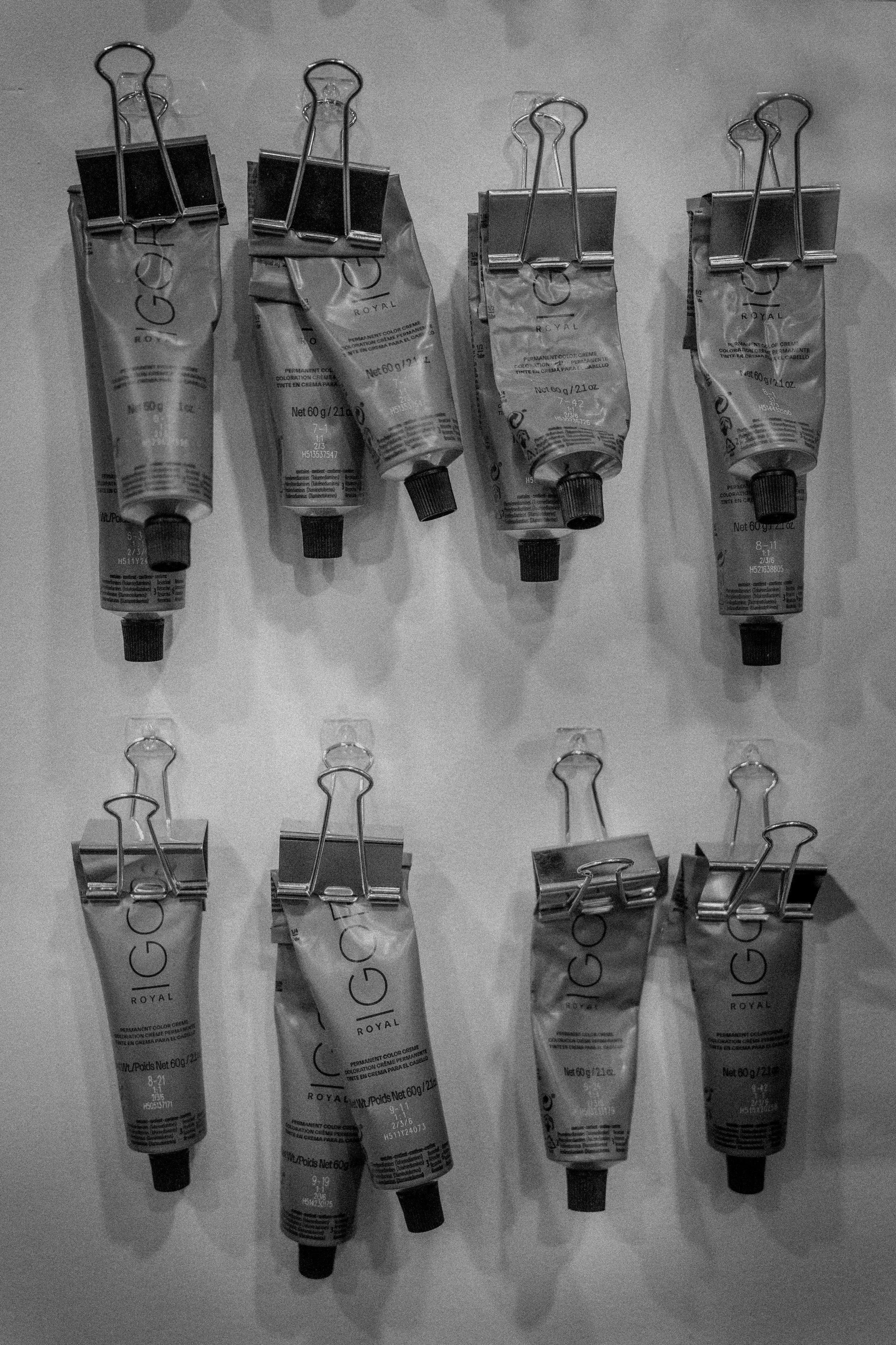Eight tubes of hair dye or cosmetic cream clipped to metal clips, arranged in two rows on a flat surface.