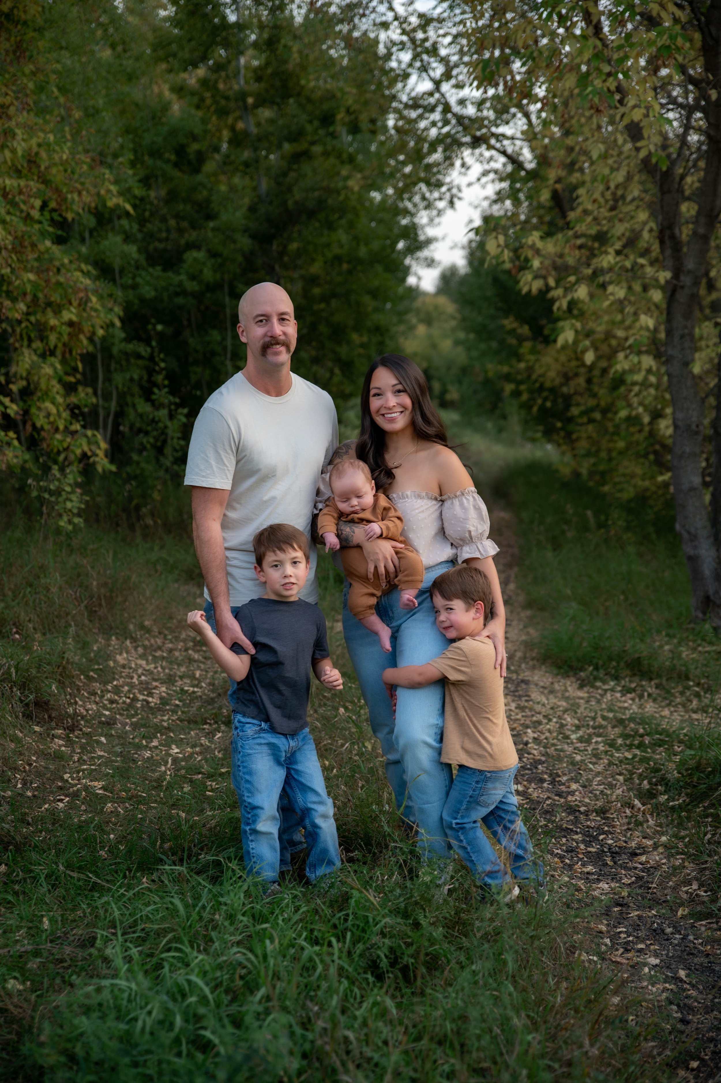 A family of five outdoors, standing on a trail surrounded by trees and greenery, smiling at the camera with a natural background during daytime.