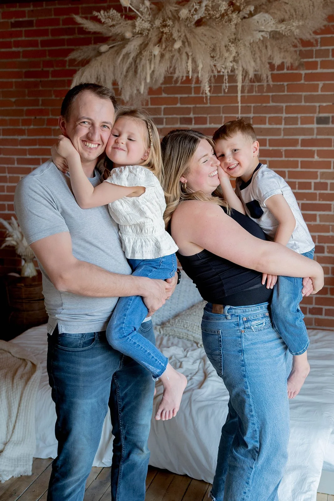 A joyful family of four with two parents and two children, posing playfully in a cozy room with a brick wall and decorative dried plants.