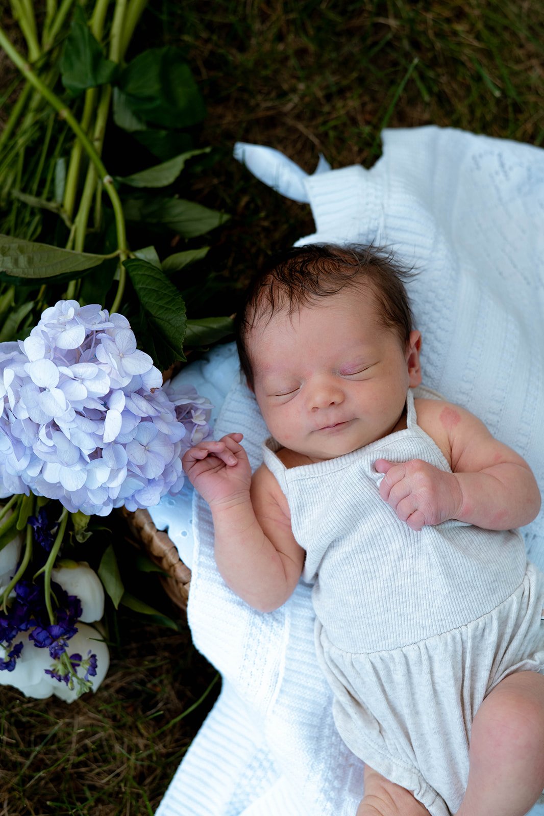 A sleeping newborn baby lying on a white blanket outdoors with purple and white flowers nearby.