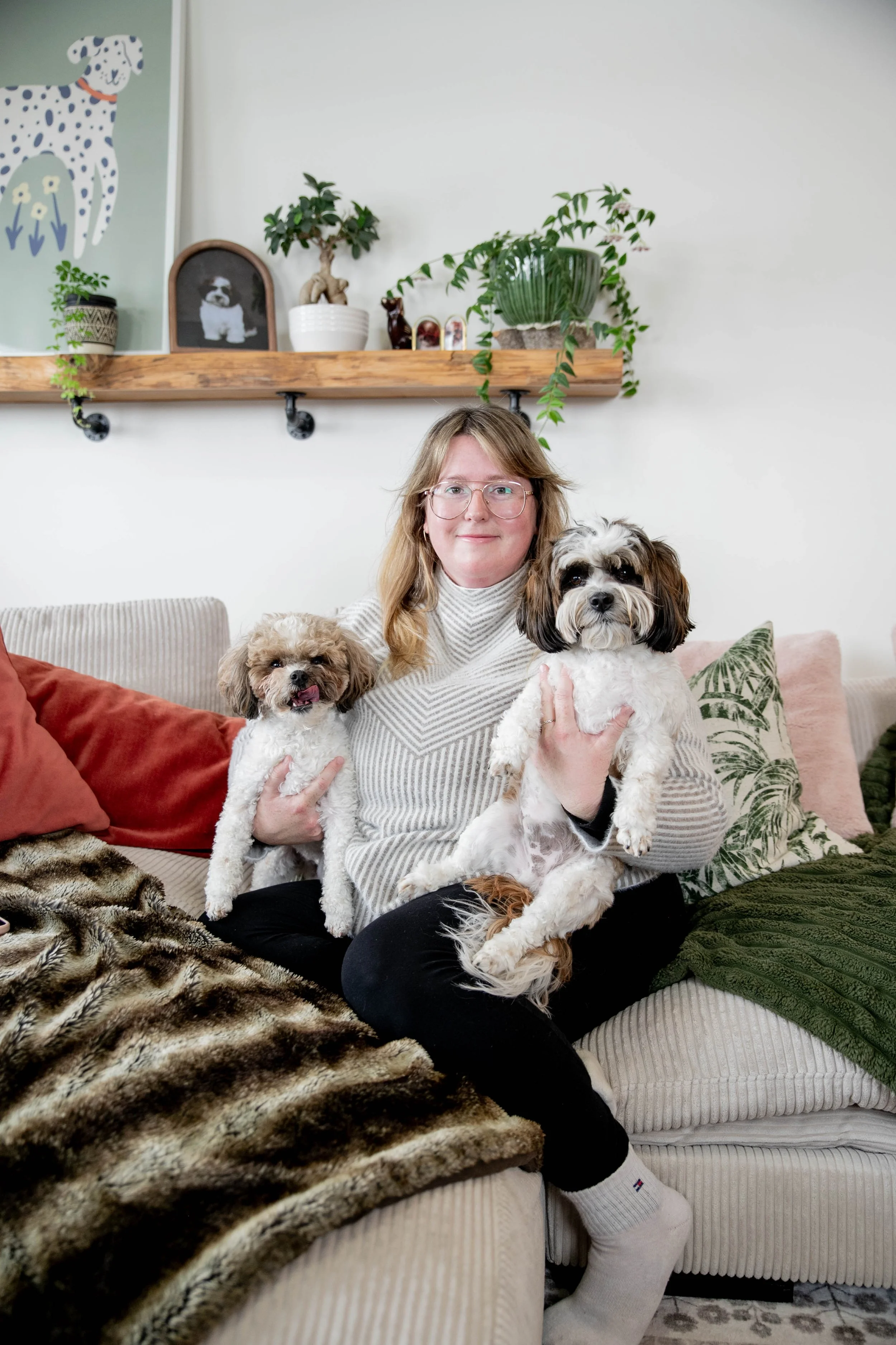 A woman sitting on a beige couch holding two small dogs, one in each arm, in a cozy living room with decorative pillows and a blanket. Behind her, there is a wooden shelf with potted plants, a framed picture of a dog, and artwork of a Dalmatian.