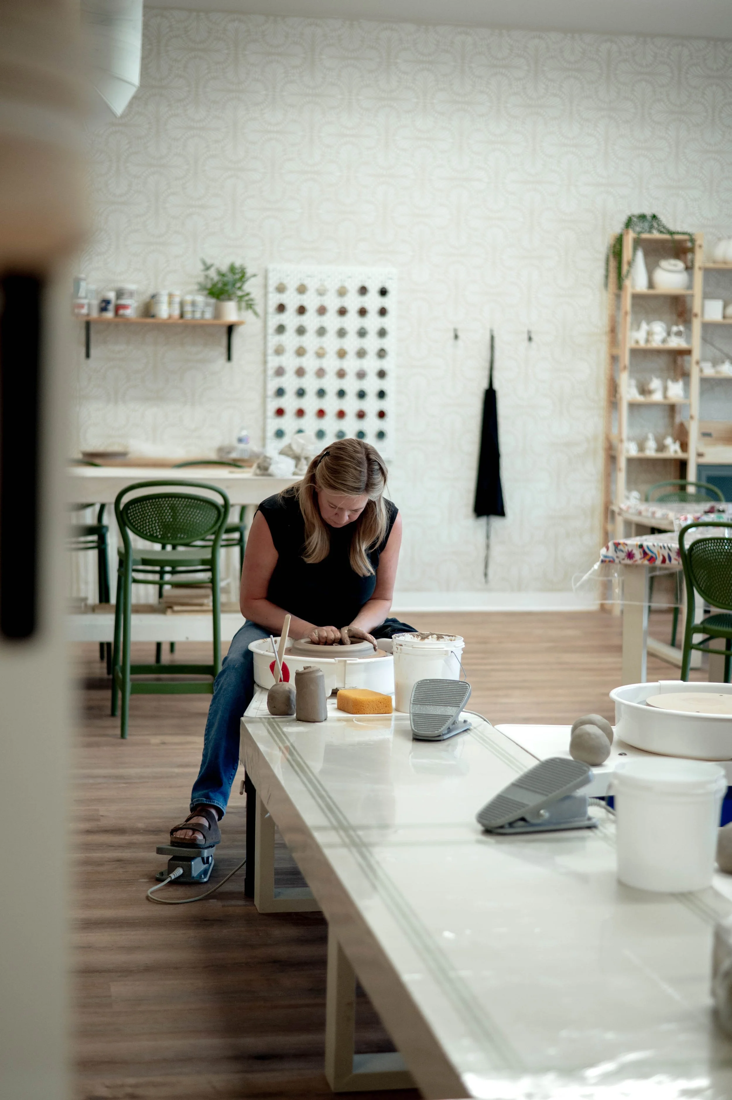 A woman working with clay on a pottery wheel in an art studio, with pottery supplies and shelves in the background.