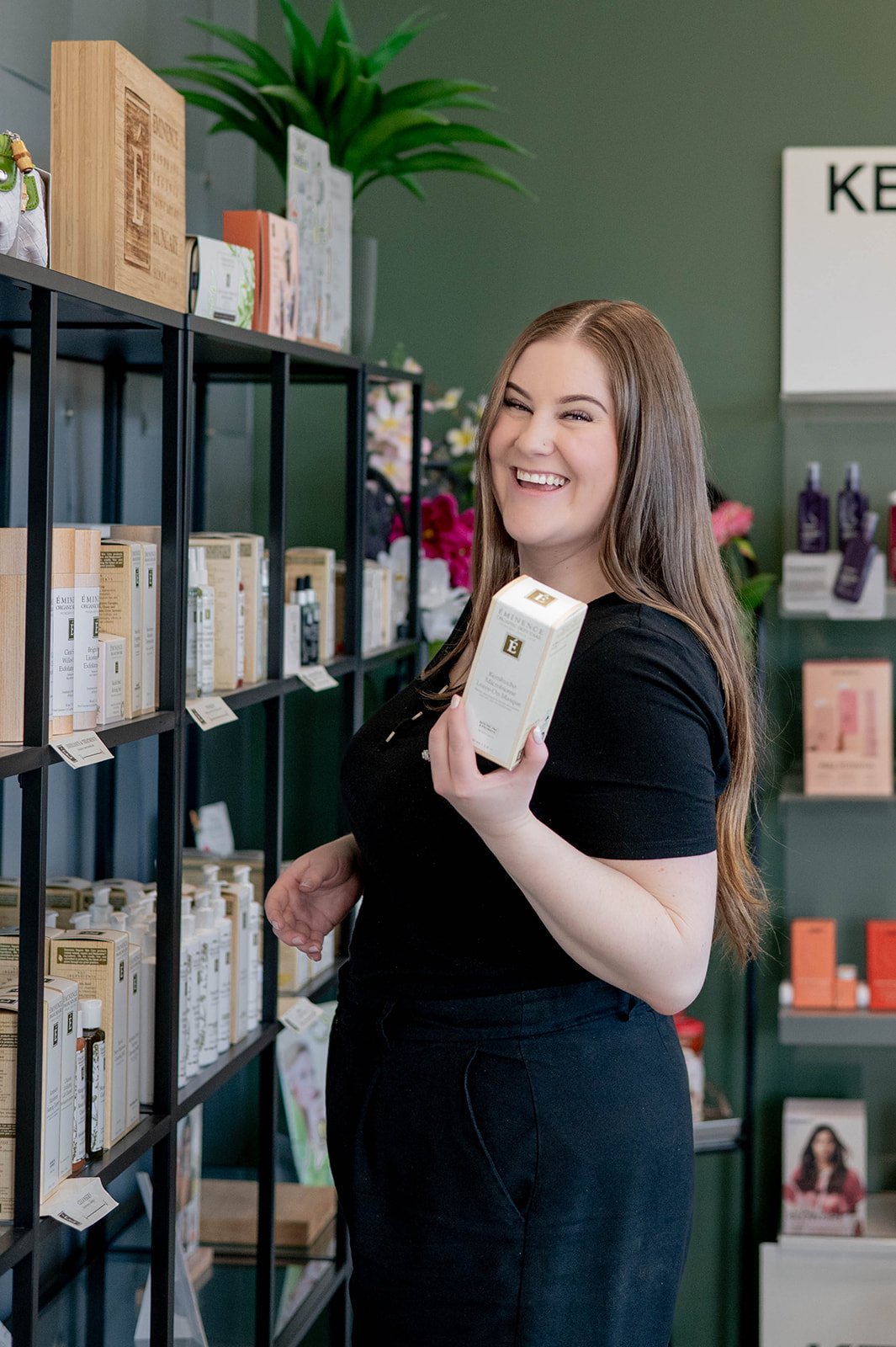 A woman with long brown hair smiling and holding a skincare product in a store with shelves filled with skincare items.