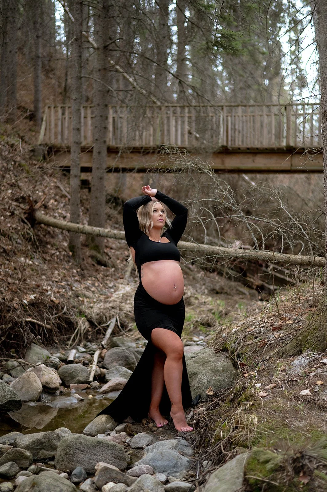 A pregnant woman in a black dress standing barefoot on rocks in a stream, with a wooden bridge in the background, in a forest setting.