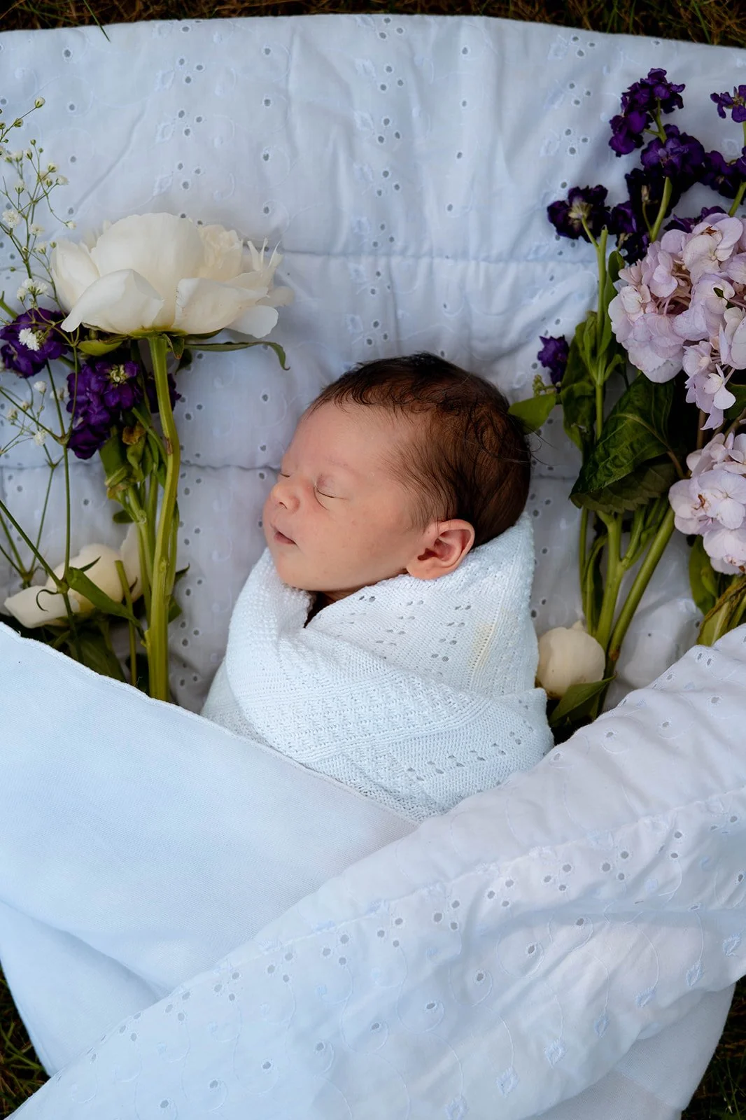A sleeping baby swaddled in a white blanket surrounded by white, purple, and pink flowers on a white embroidered cloth.