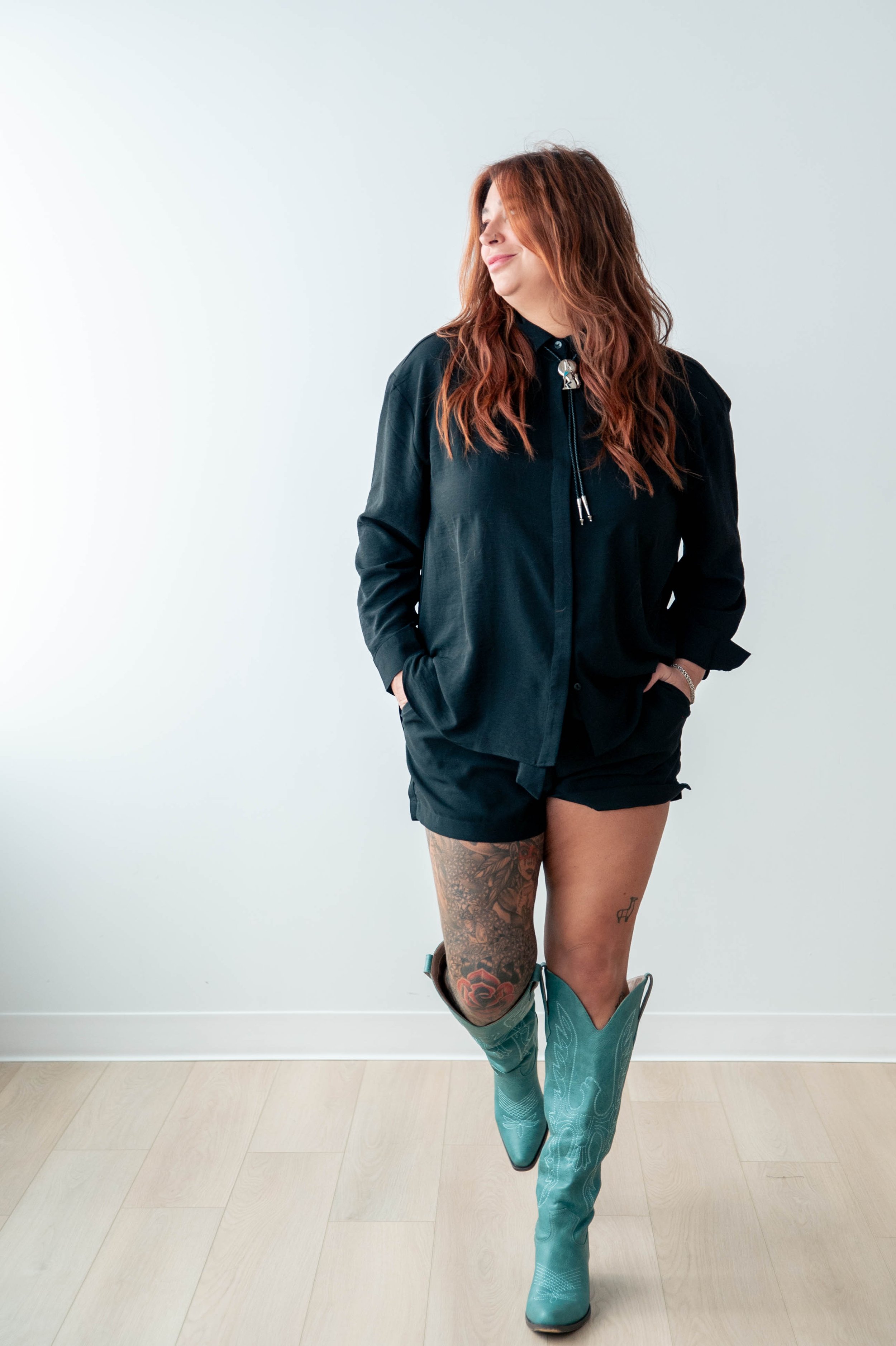 A woman with long wavy red hair wearing a black zip-up jacket, shorts, turquoise cowboy boots, and showing tattoos on her left thigh.