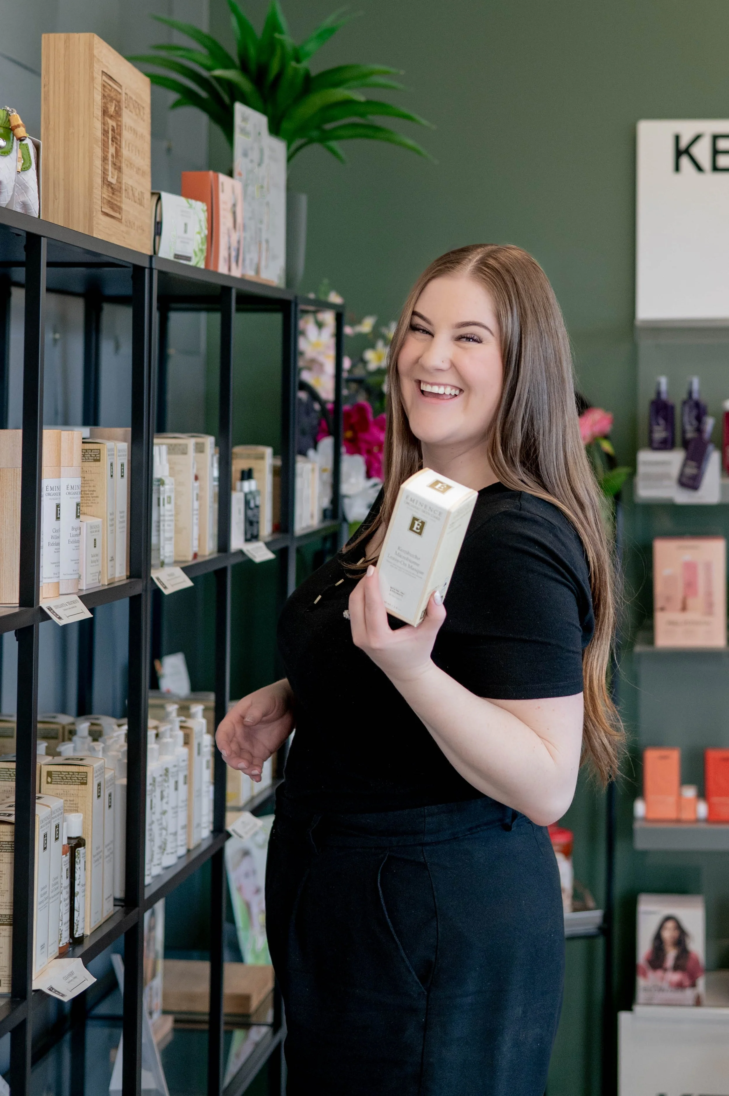 A woman with long brown hair smiling and holding a box of skincare product inside a store with shelves of skincare products and a green wall background.