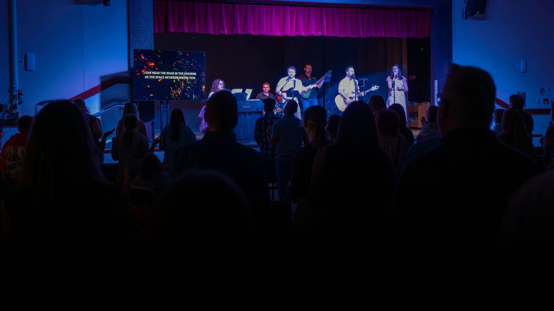 A singer and a group of musicians perform on a stage in front of an audience. There is a screen displaying lyrics and a pink curtain hanging above the stage.