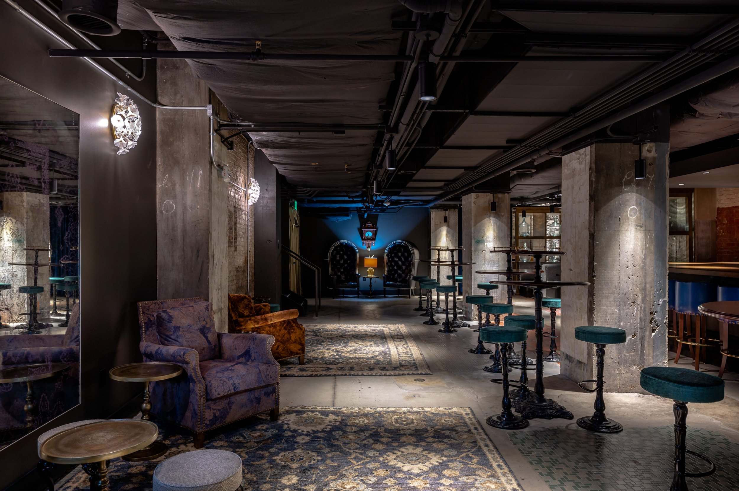 Empty bar with vintage decor, velvet armchairs, high-top tables, patterned rugs, concrete pillars, and dim lighting.