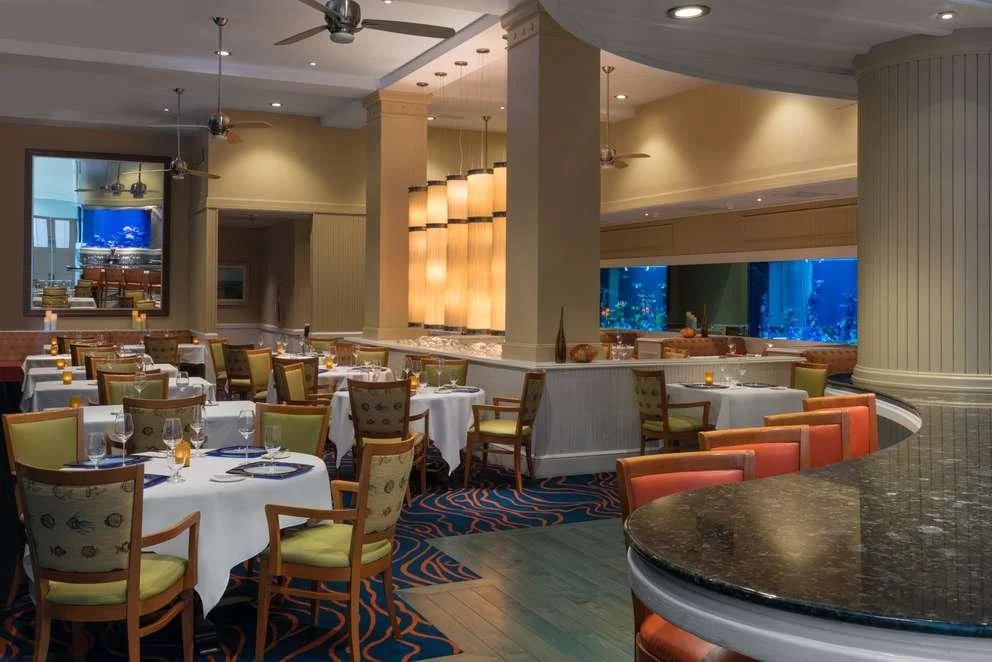 Elegant restaurant interior with round tables covered in white tablecloths, set with glassware and utensils, surrounded by varied colorful chairs, large aquarium windows, and decorative lighting.