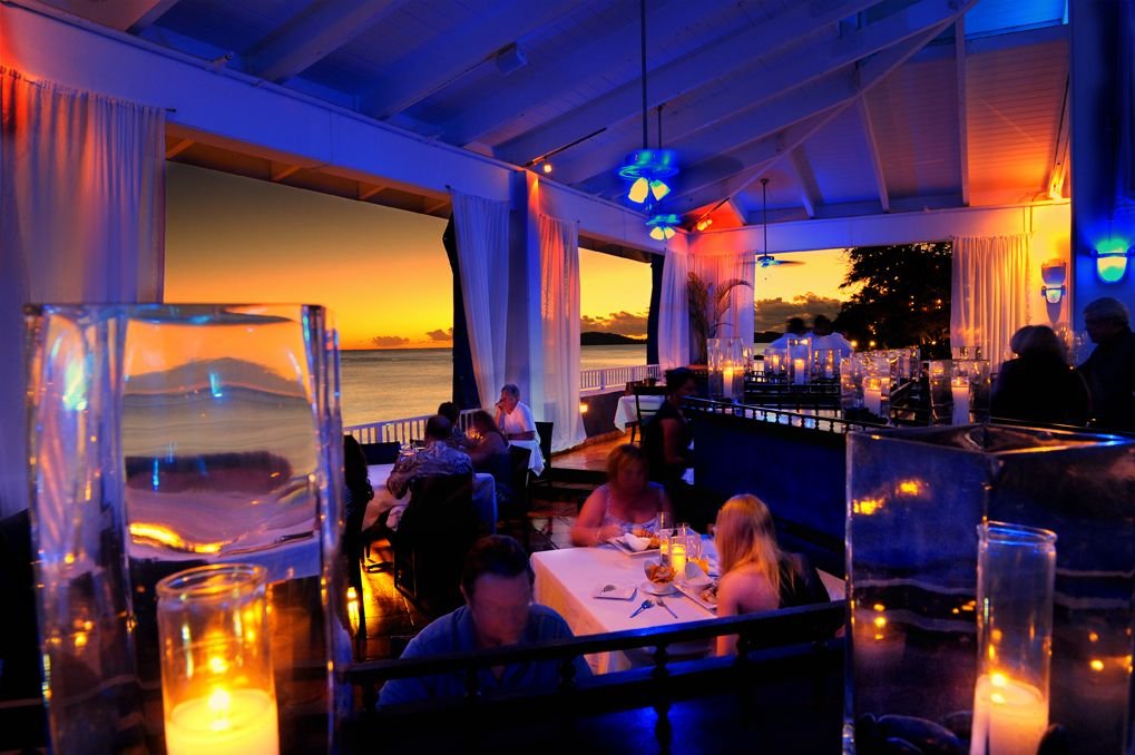 A restaurant with open sides overlooking the ocean at sunset. The interior is decorated with candles and colorful ceiling lights, and diners sit at tables enjoying the view.