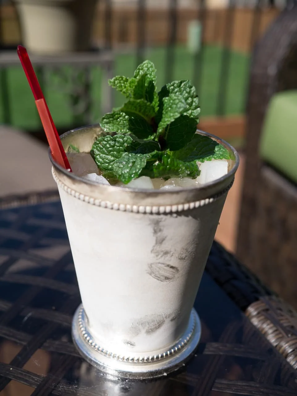A close-up of a glass with a mint garnish on top, containing a cold drink with ice, on an outdoor table with a blurred background.