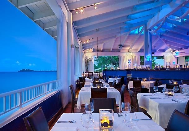Elegant waterfront restaurant interior with white tables, black chairs, candlelit table settings, and large windows overlooking the ocean during dusk.