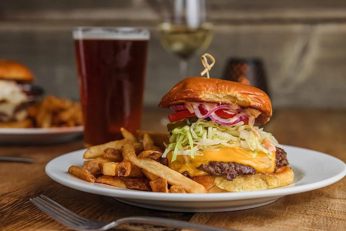 A cheeseburger with lettuce, tomato, onions, and cheese on a bun, served with French fries and a glass of dark soda on a wooden table.