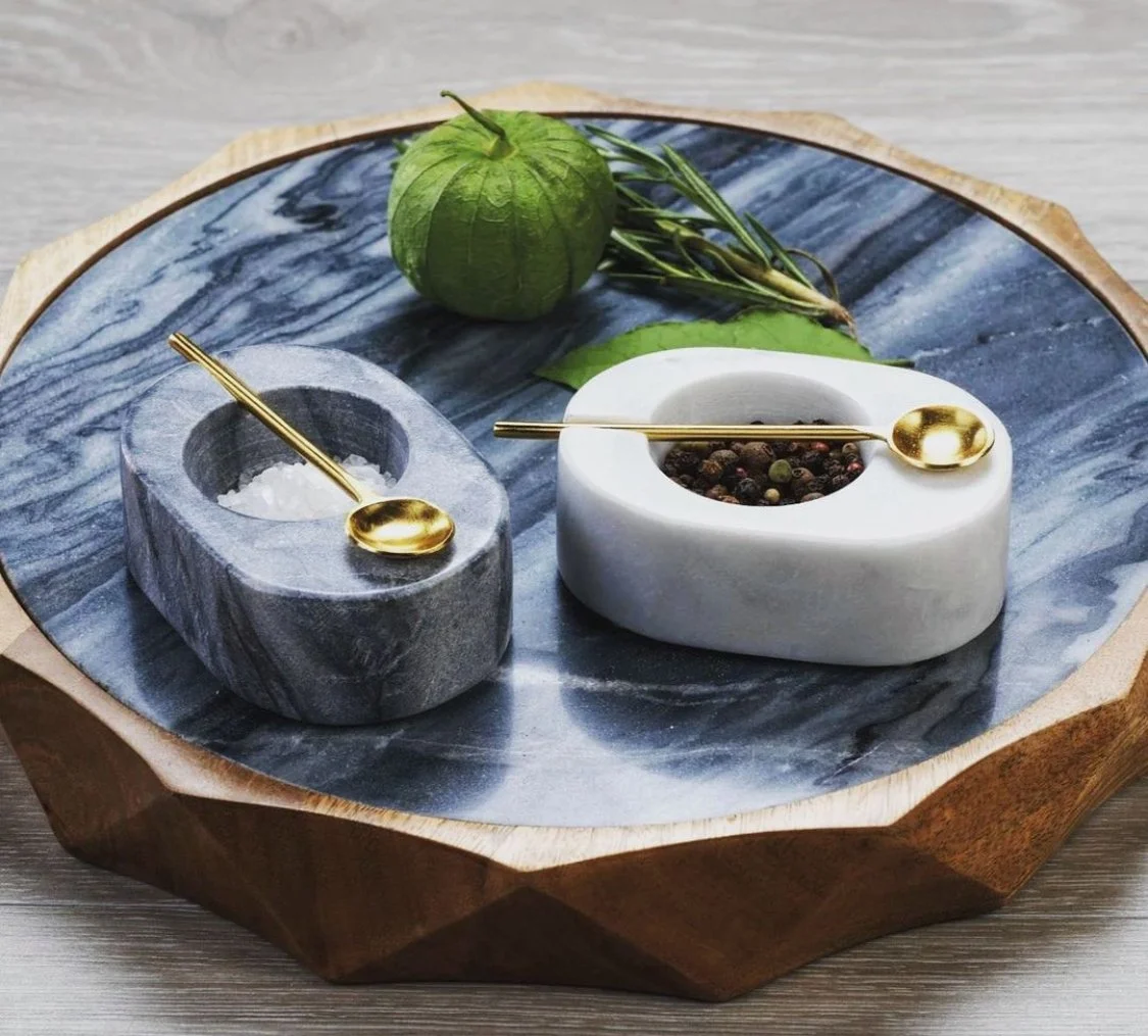A marble and stone salt and pepper set with gold spoons on a round marble tray, with a small green fruit and sprigs of herbs in the background.