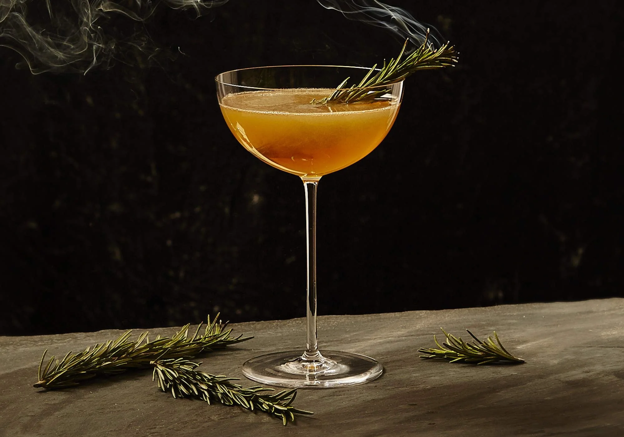 A cocktail garnished with a sprig of rosemary in a coupe glass, with additional rosemary sprigs on a gray surface in front of a black background.