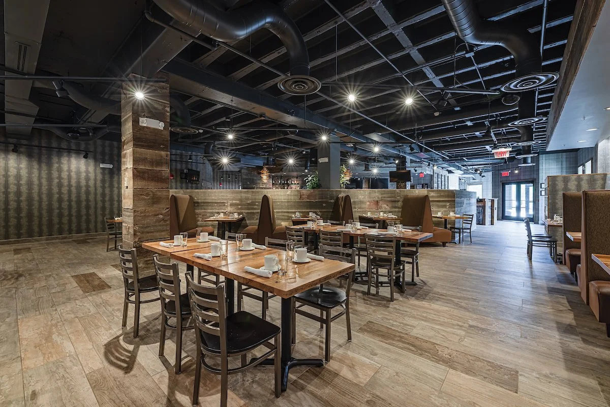 Empty restaurant dining area with wooden tables and chairs, some booths with high backs, modern industrial ceiling with black ducts and lighting fixtures, near entrance with glass doors.